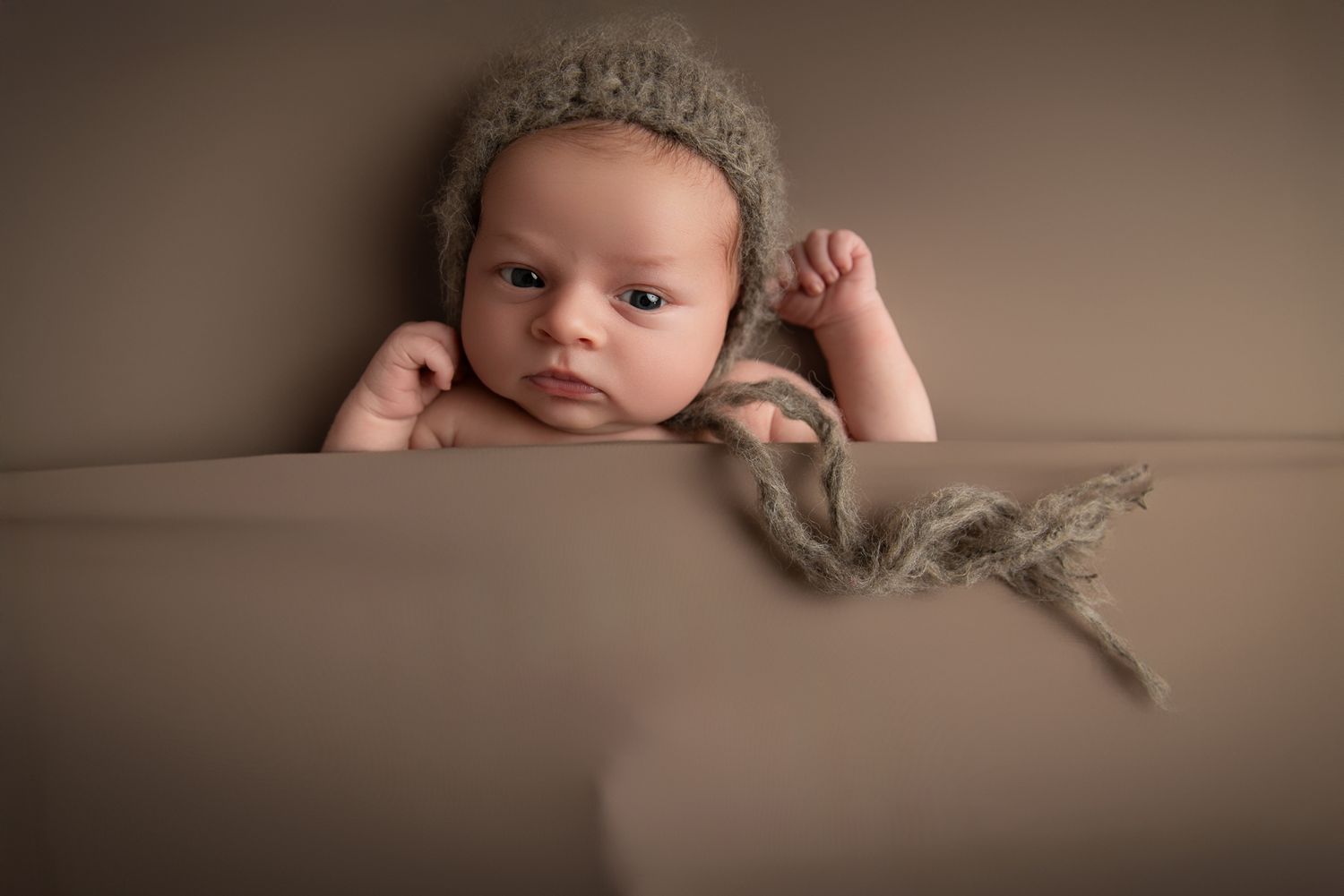 Alert newborn baby in knit gray bonnet peeking over edge of soft brown blanket with curious eyes. Fallbrook Newborn Photographer
