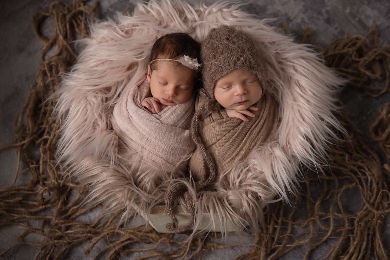 Twin newborns swaddled in neutral wraps sleep together in a fluffy fur nest with rope accents in Oceanside