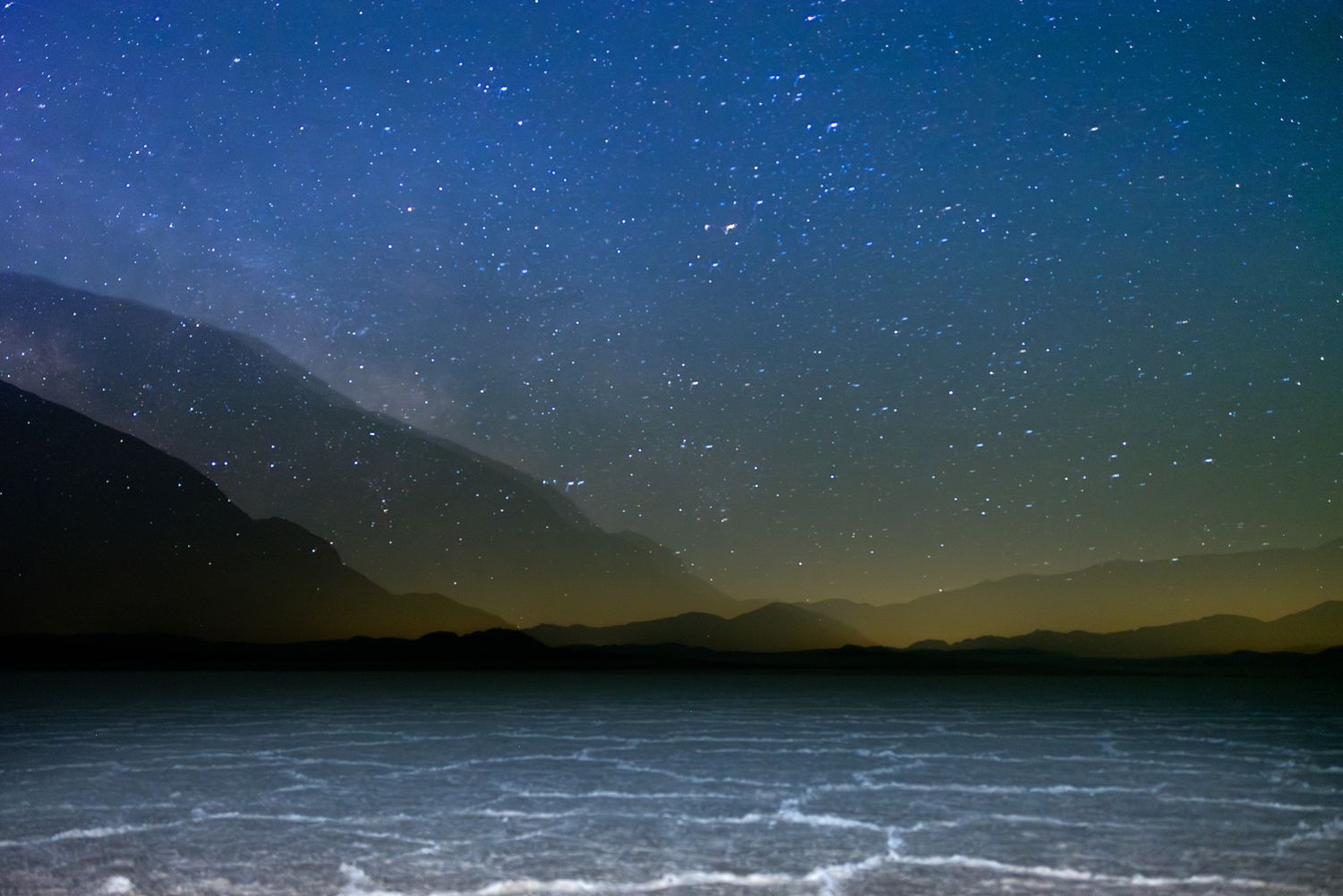 Starry night sky over silhouetted mountains reflecting on salt flats in a serene desert landscape.