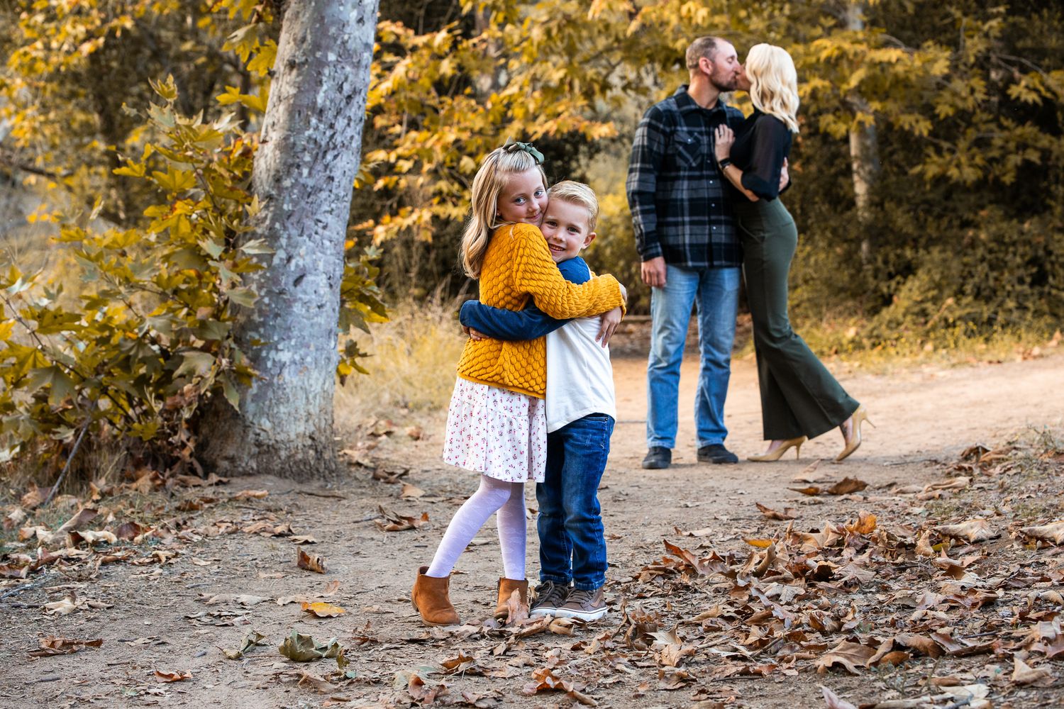 Two young siblings hug on a leaf-covered path while parents kiss in the background.