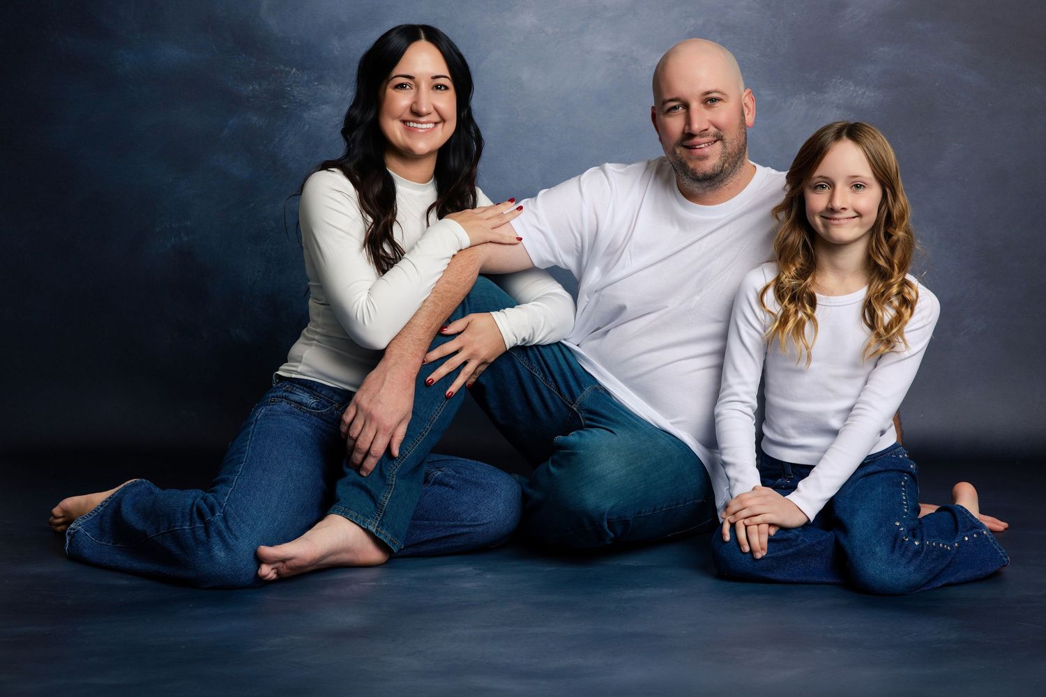 Family of three in matching white tops and jeans posing together against a blue-gray studio backdrop.