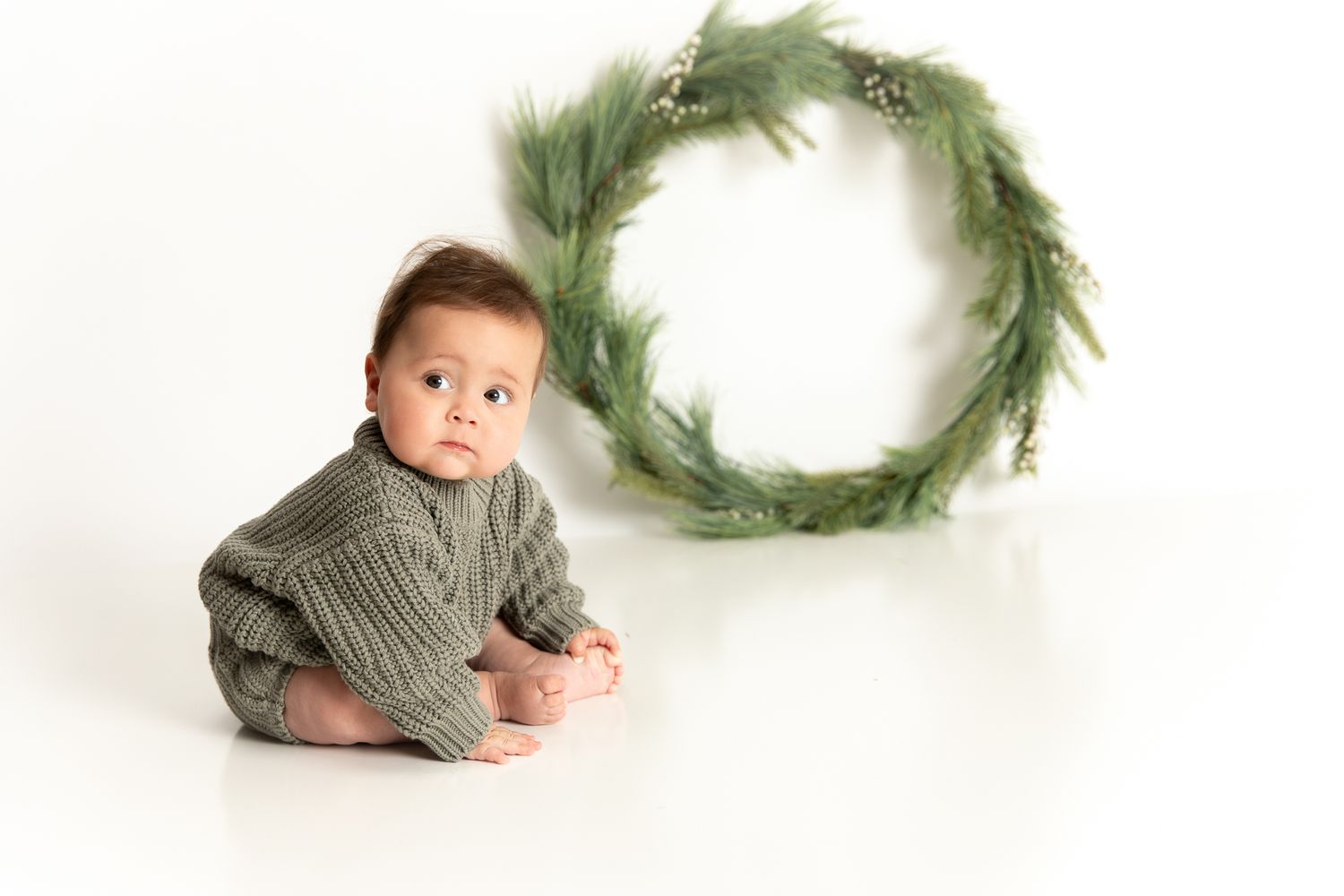 Baby in green knit outfit sits on white floor beside a festive evergreen wreath for a holiday photo.