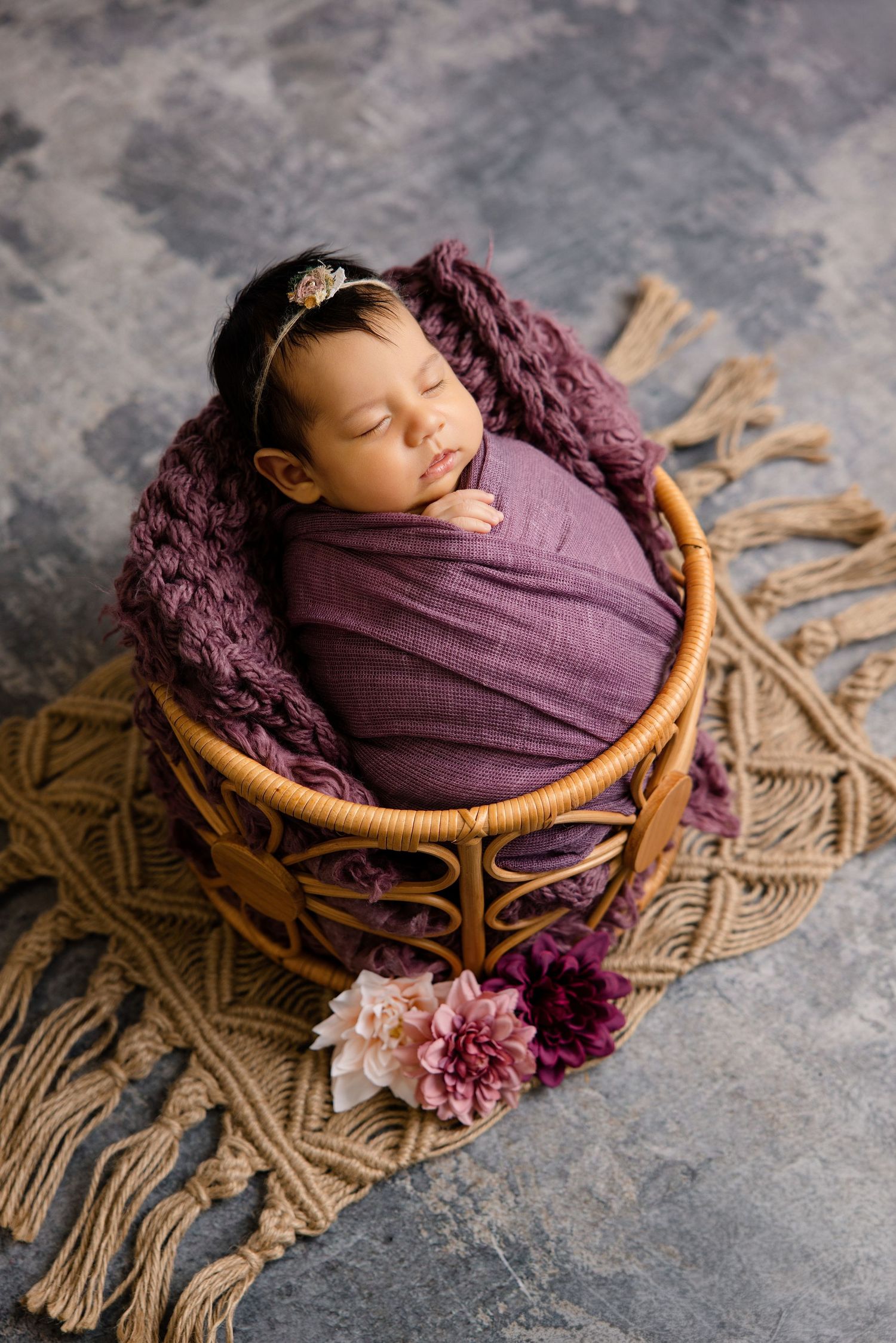 Peaceful newborn baby swaddled in purple wrap resting in a wicker basket with flowers.