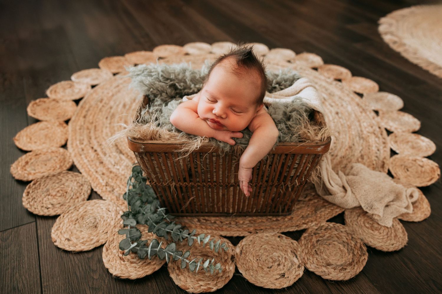 Sleeping newborn baby swaddled in gray wrap resting in a wicker basket on a round jute rug.