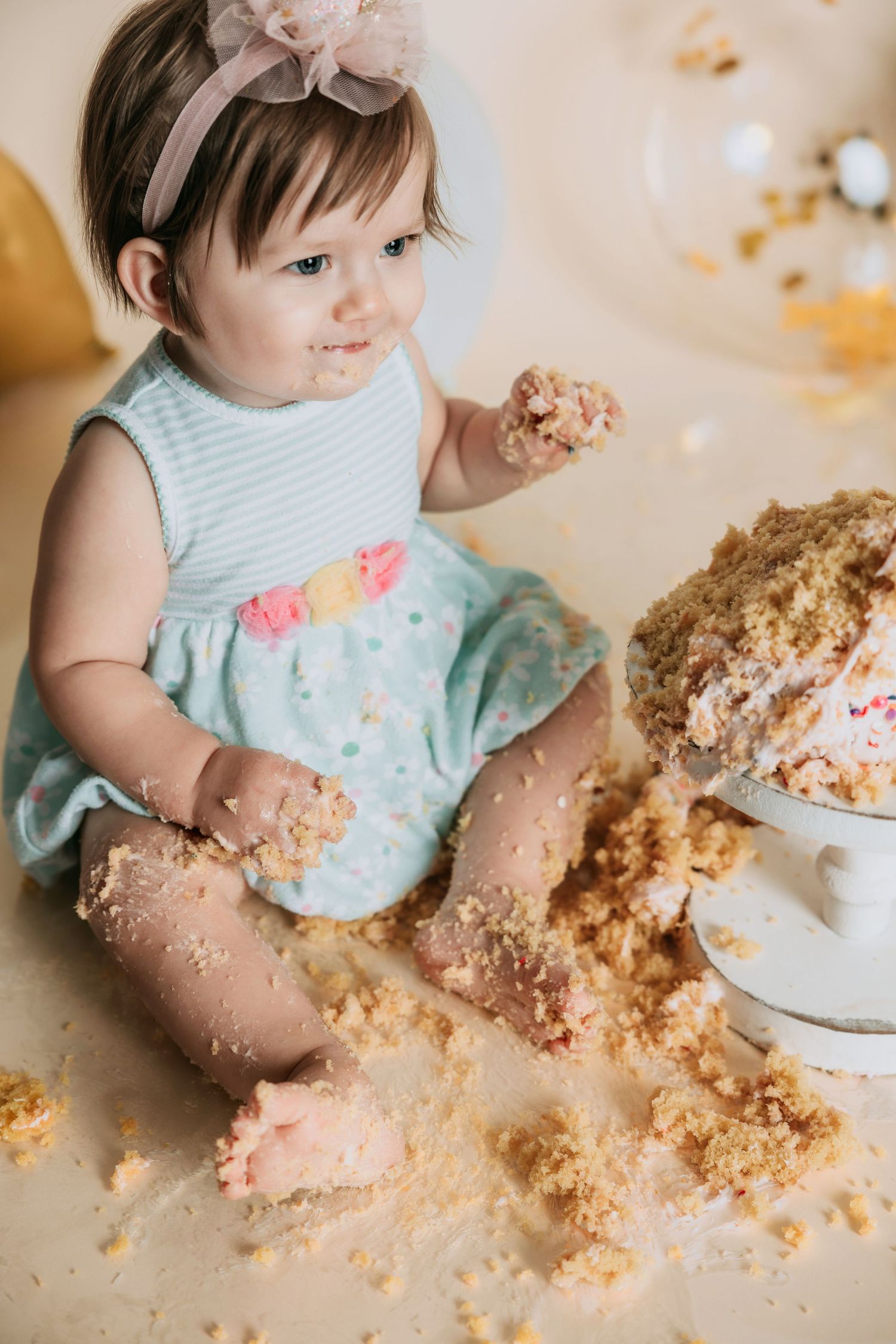 Cute baby girl in mint dress smashing first birthday cake, covered in crumbs, sitting on floor with balloons.