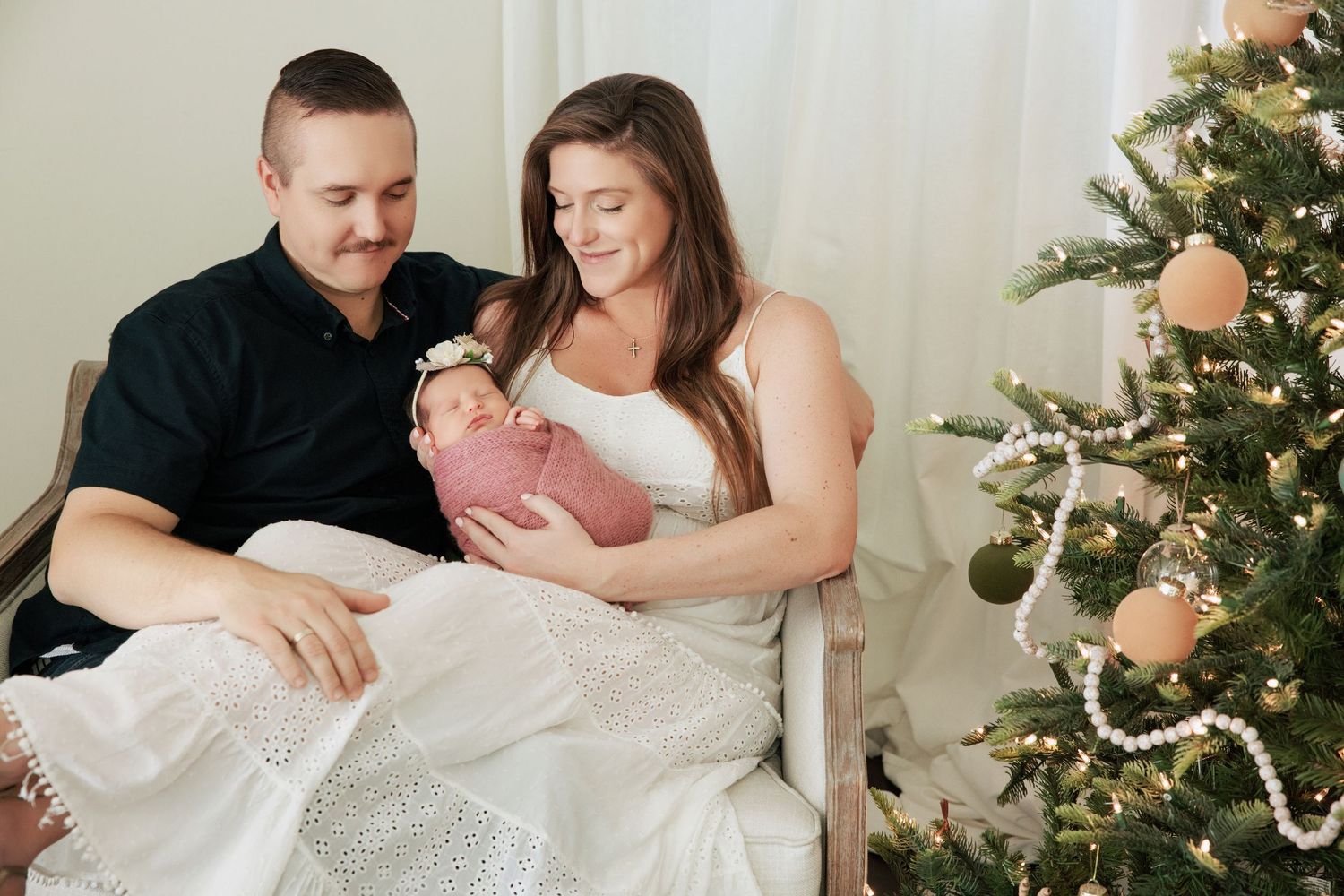 A couple sits together holding their newborn baby wrapped in pink near a decorated Christmas tree.