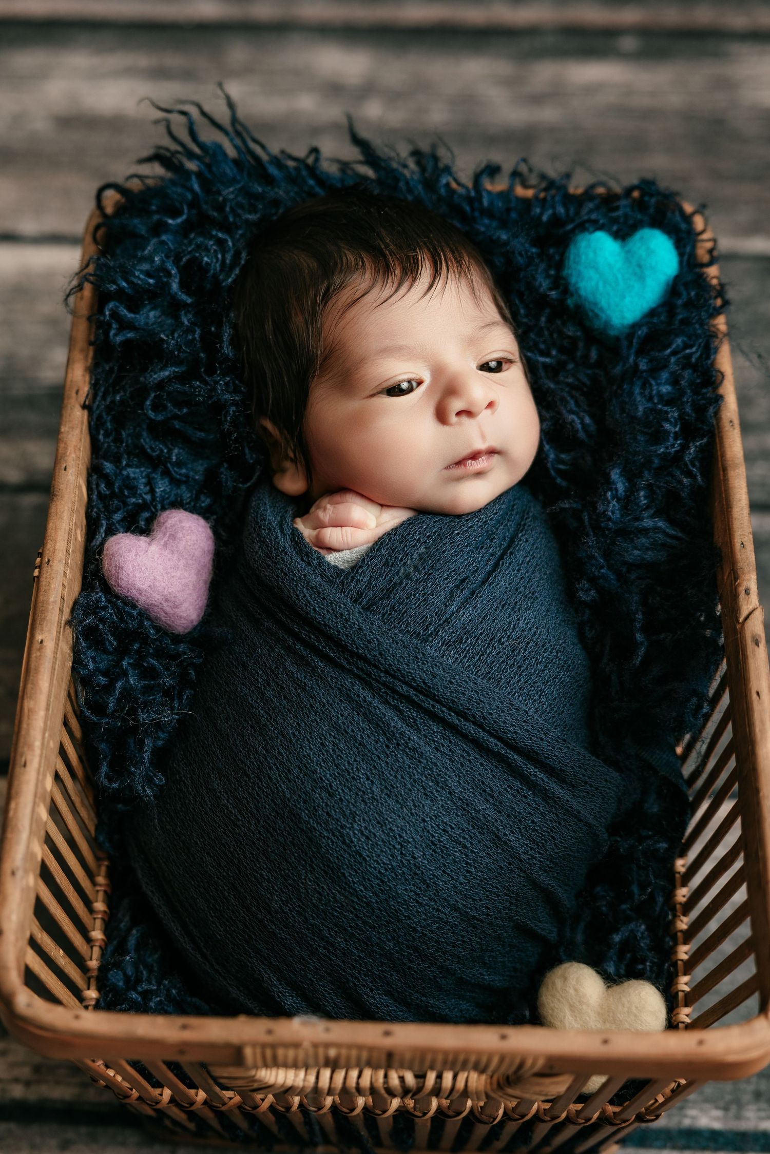 Awake newborn baby wrapped in dark blue swaddle in wicker basket with colorful felt hearts.