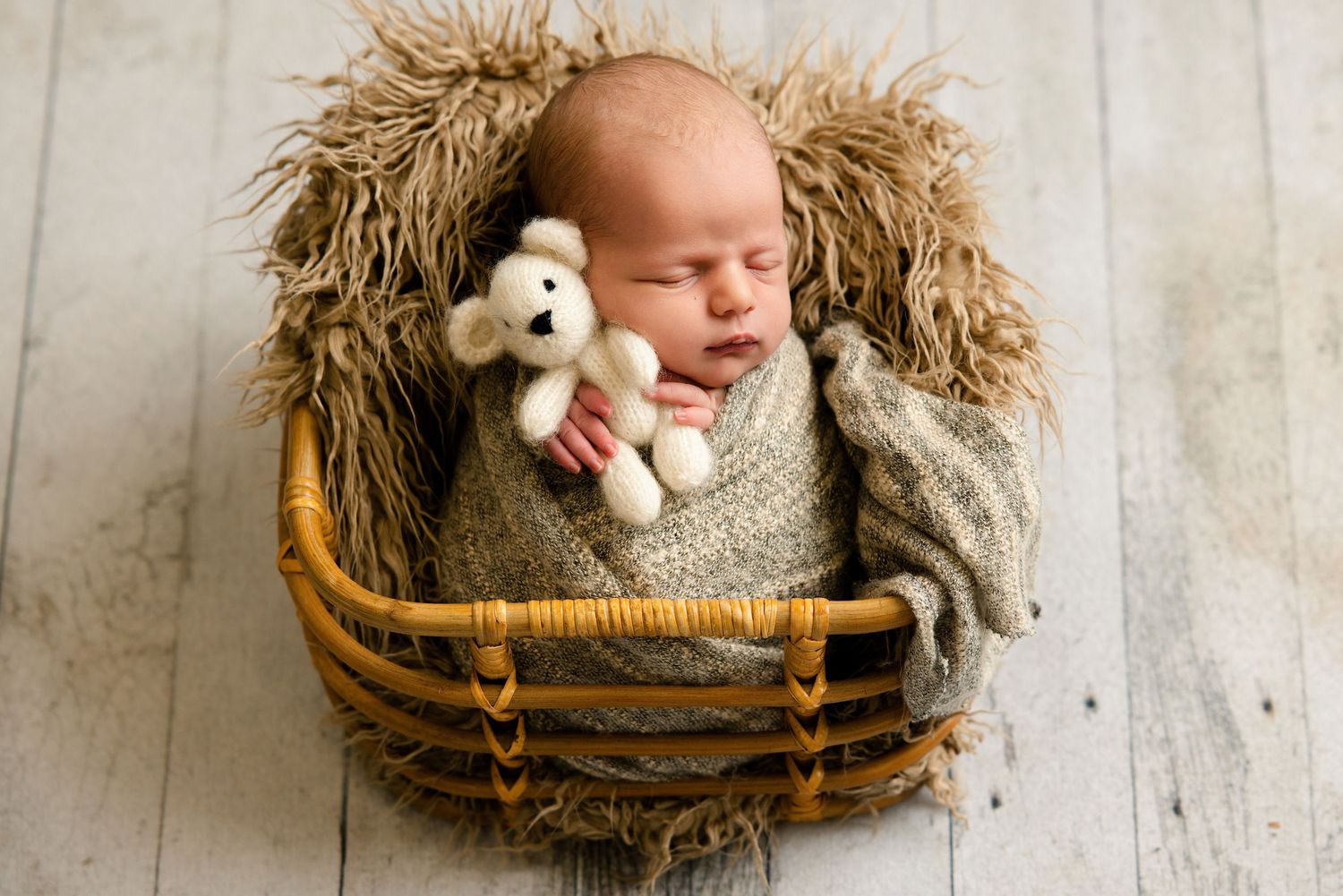 Newborn baby sleeping in a wicker basket wrapped in a knit blanket, holding a small white teddy bear.