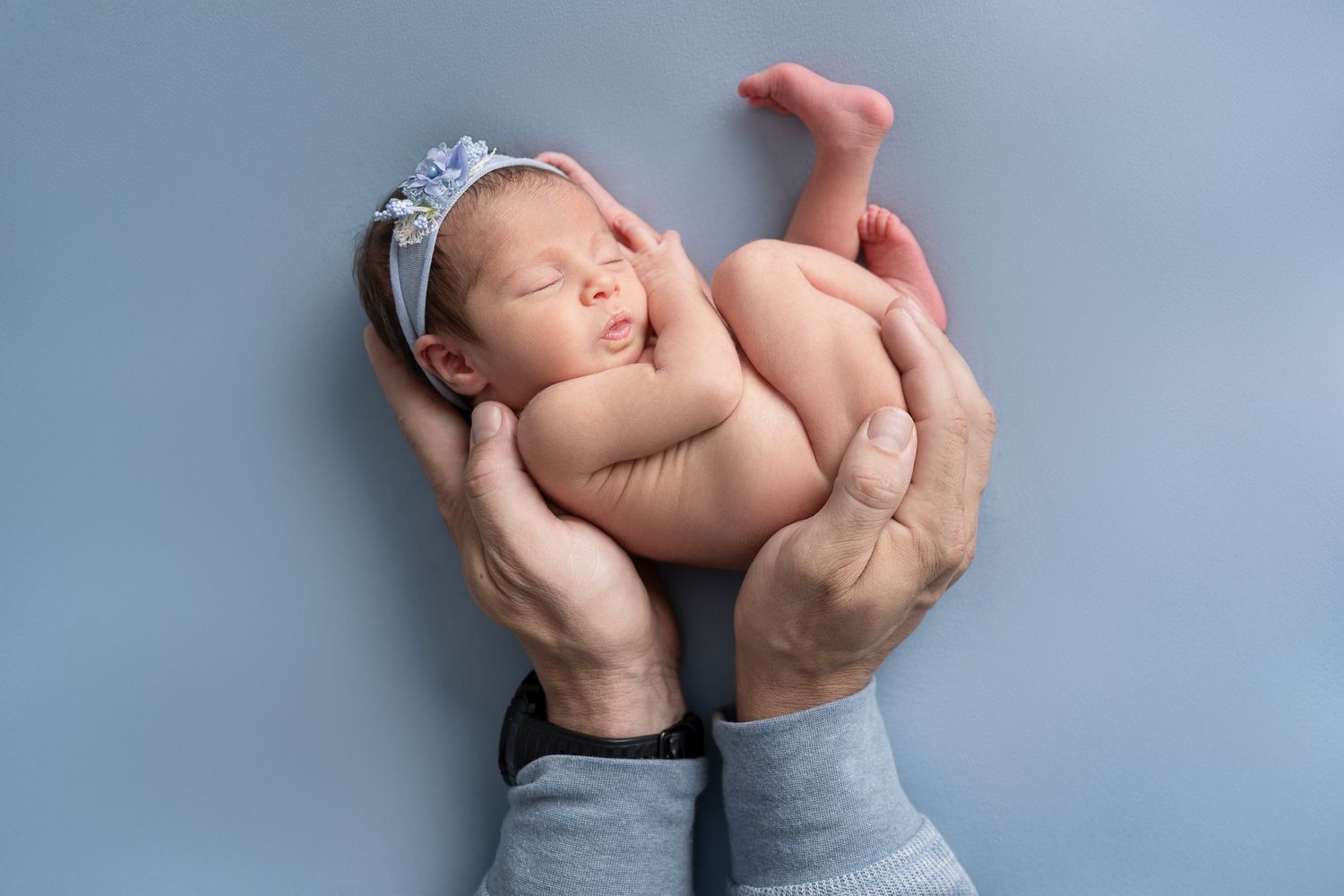 Newborn baby girl with floral headband sleeping peacefully in parents hands against soft blue background.