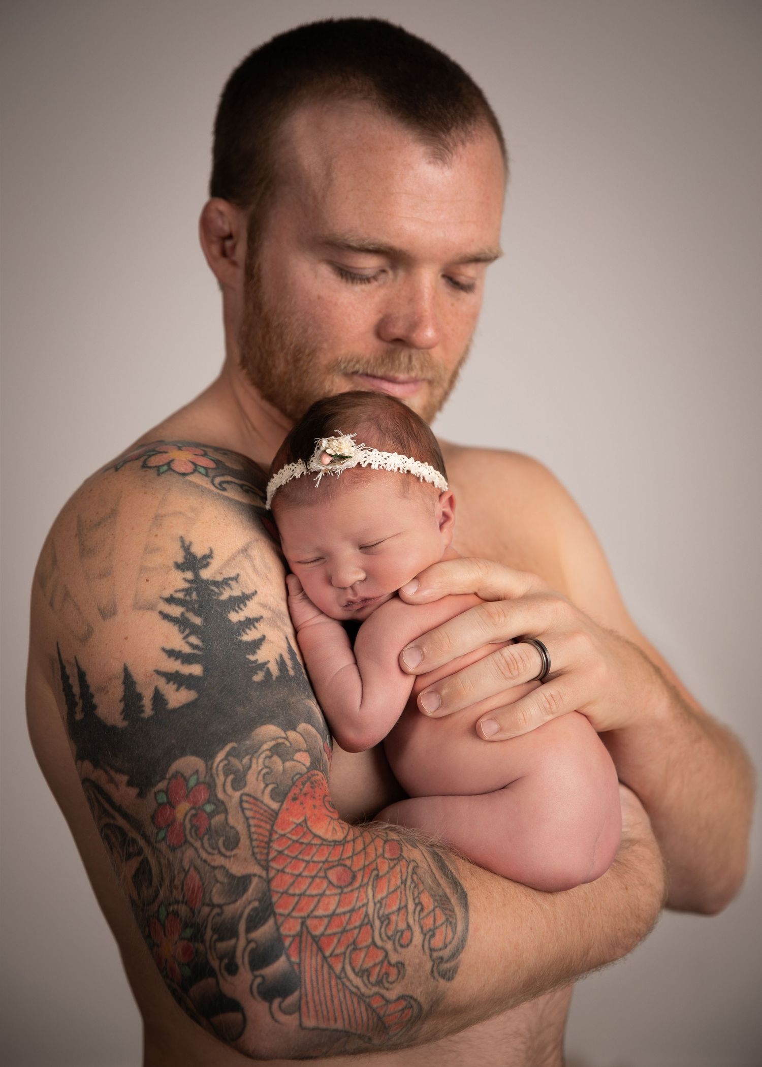 Tattooed father tenderly holds sleeping newborn baby girl wearing white headband against his chest.