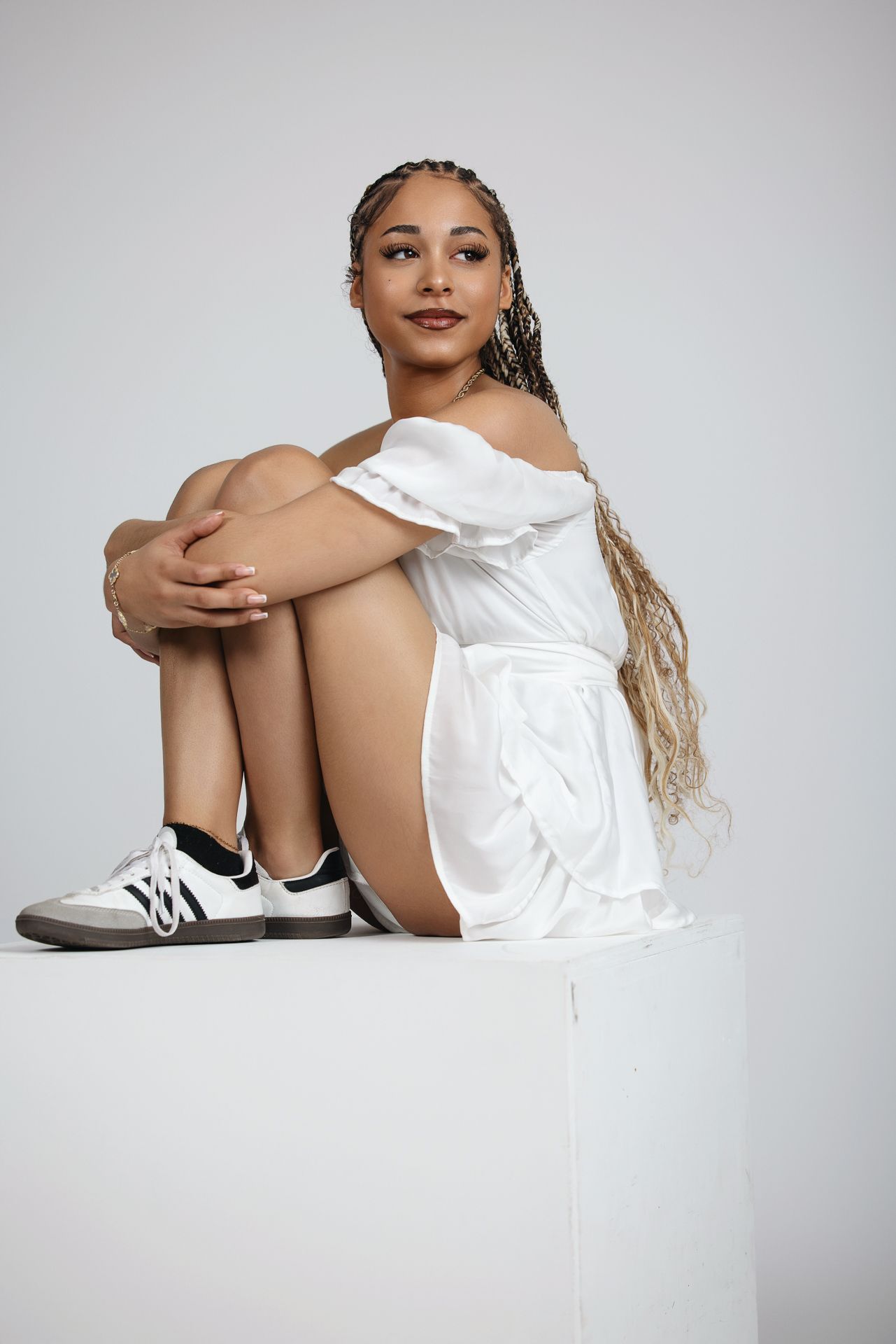 Young woman with braided hair in white off-shoulder dress sitting on white cube in studio portrait. Senior photography session in Bonasll ne