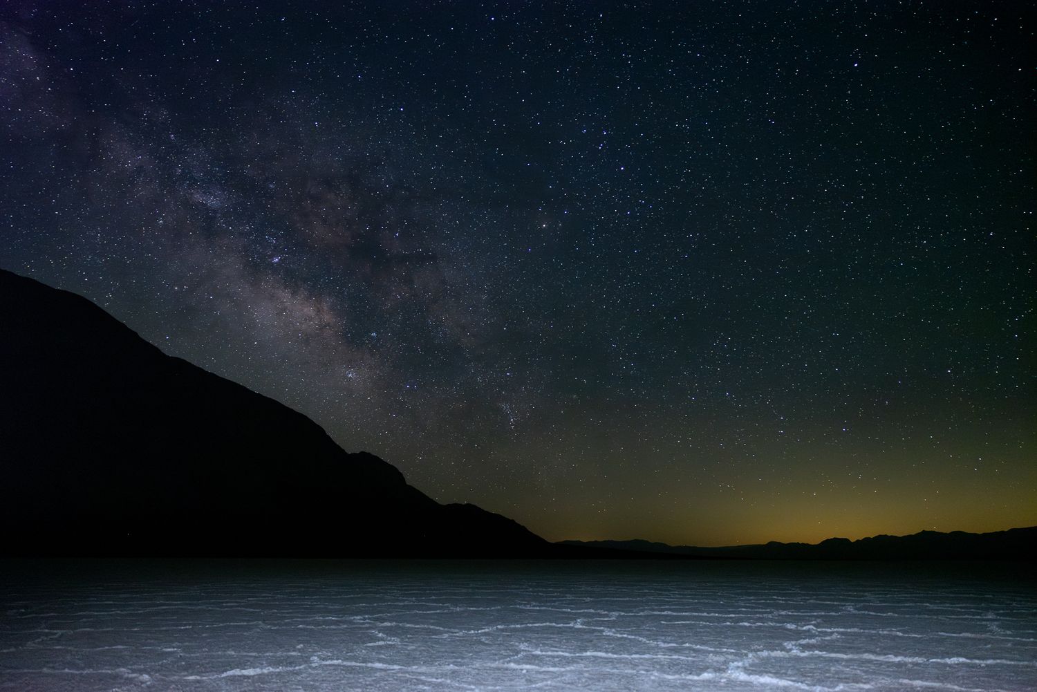 Milky Way galaxy glowing over dark desert salt flats at night with silhouetted mountains and golden horizon light.