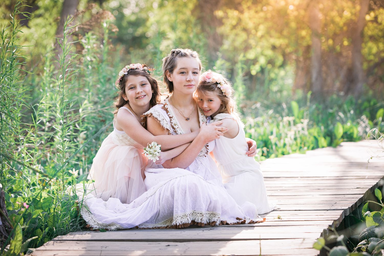 Three girls in white dresses with floral crowns sit together on a wooden bridge in a lush garden.