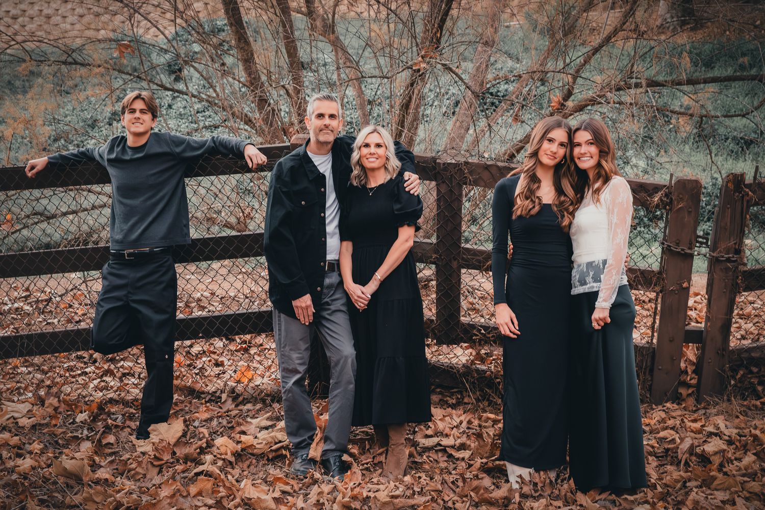 Family of five posing by a wooden fence surrounded by autumn leaves in an outdoor fall photo session.