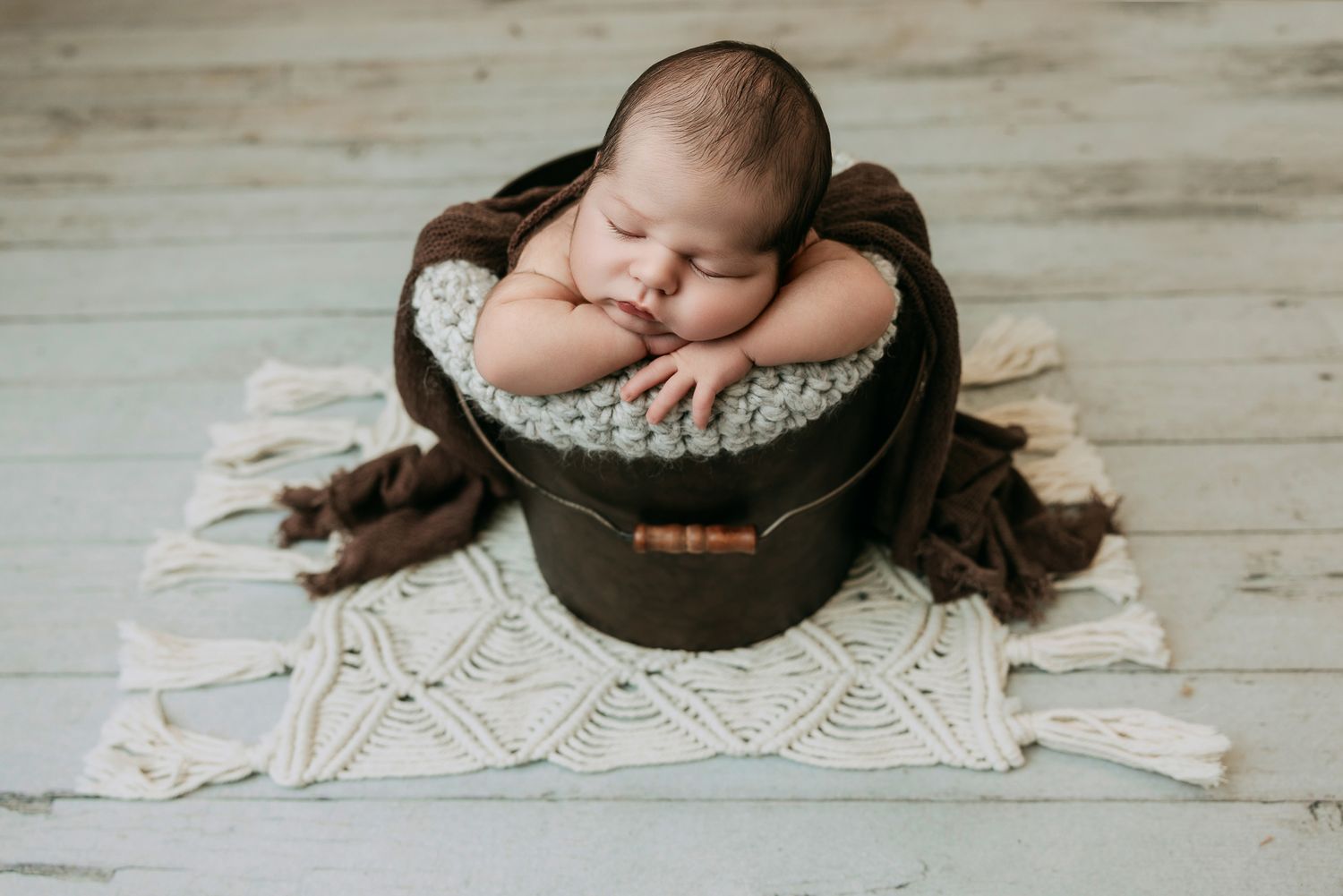Sleeping newborn baby rests chin on hands inside a dark wooden bucket on a macrame rug.