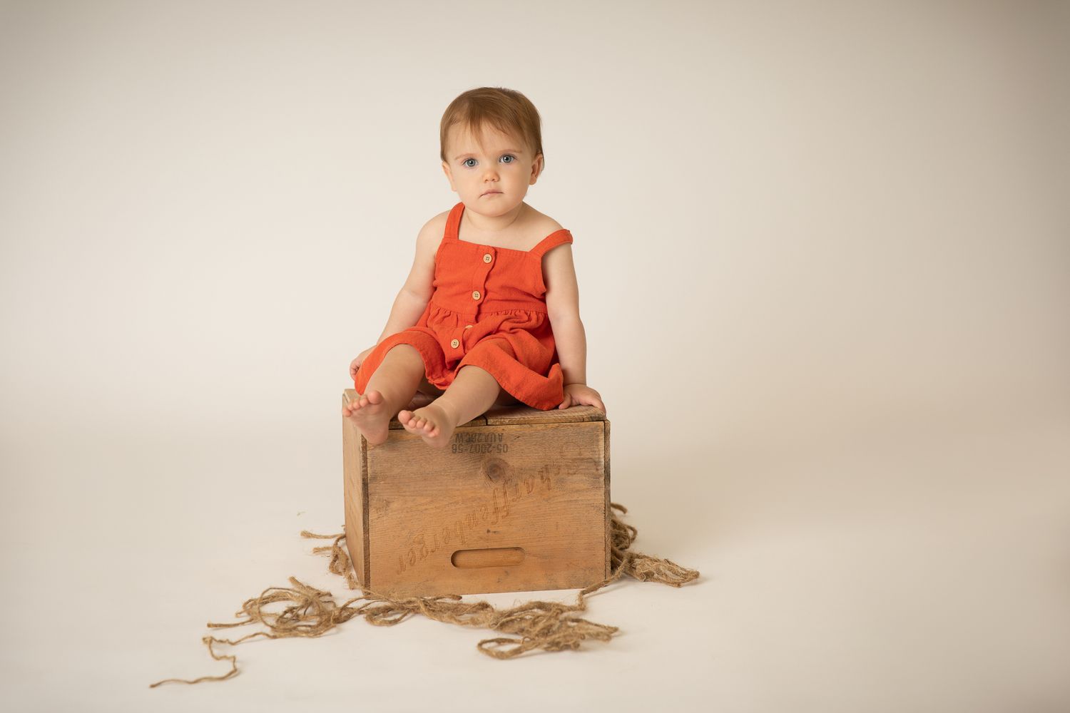 Cute toddler in red romper sitting on rustic wooden crate with rope.