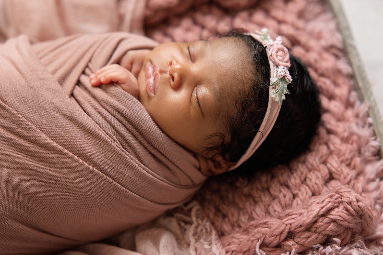 Newborn baby girl swaddled in pink wrap wearing floral headband, sleeping peacefully in a cozy knit blanket.