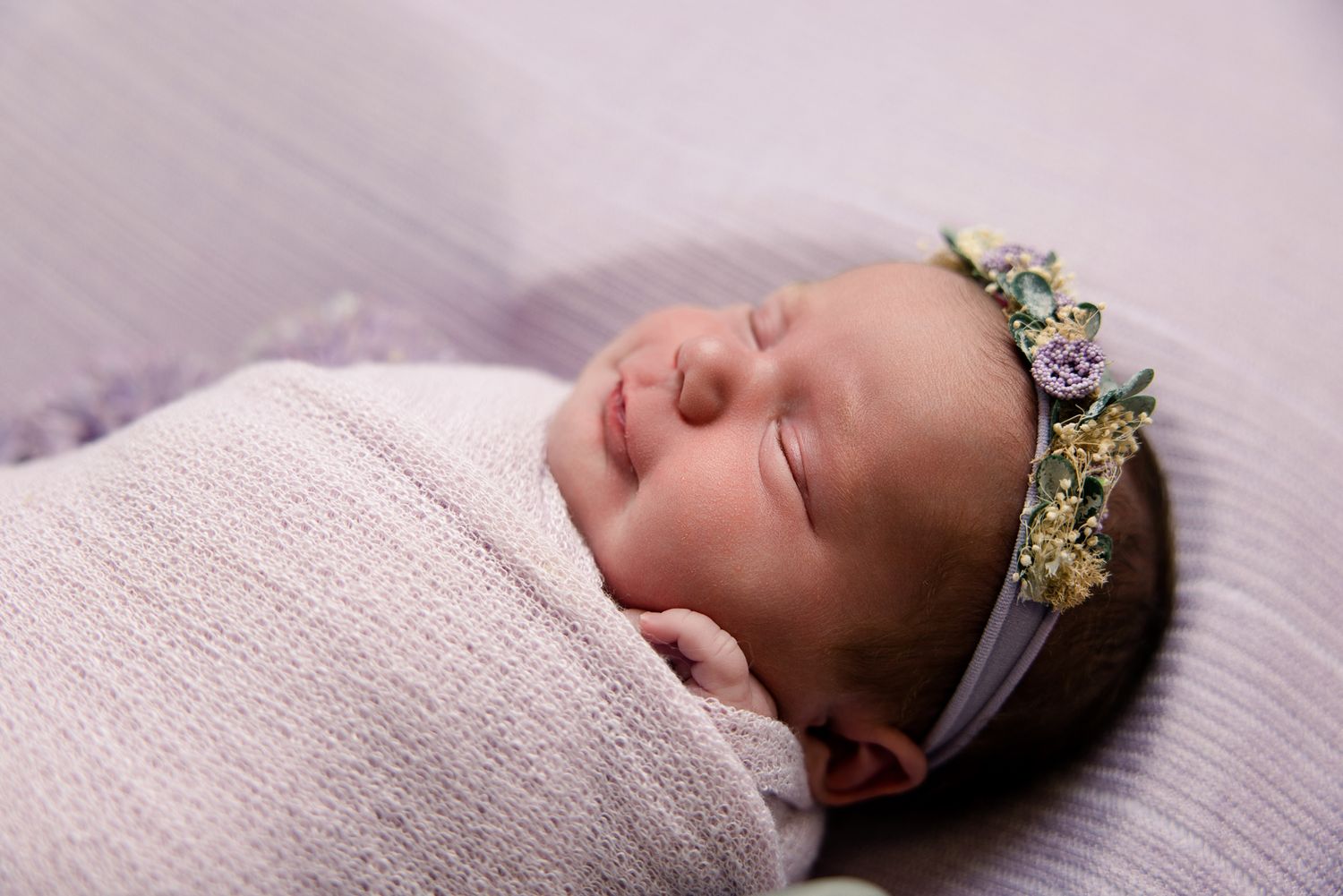 Peaceful newborn baby girl sleeping swaddled in white wrap, wearing a delicate floral headband.