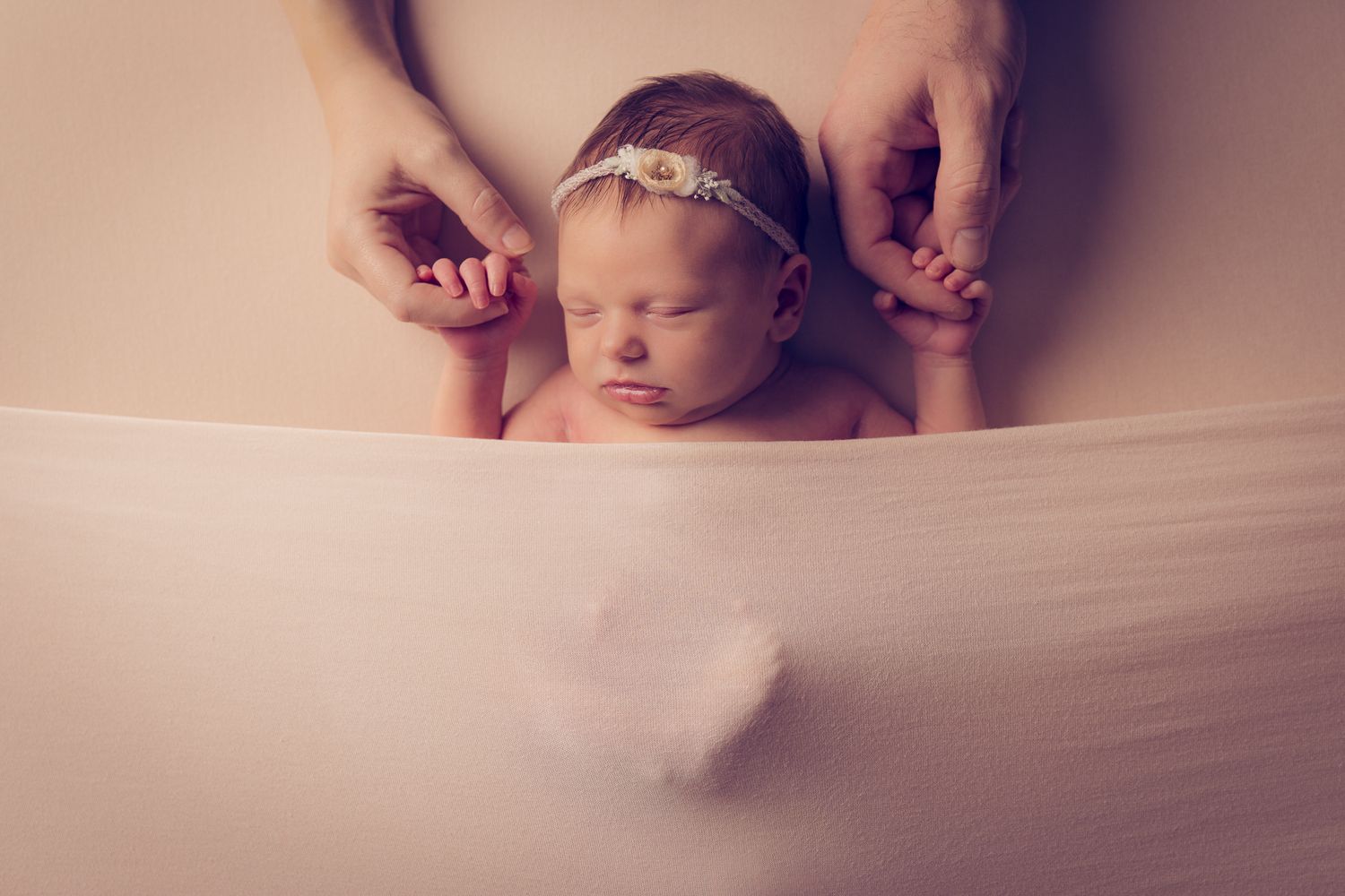 Peaceful newborn baby girl sleeping on soft pink fabric, held gently by parents hands, wearing delicate floral headband.