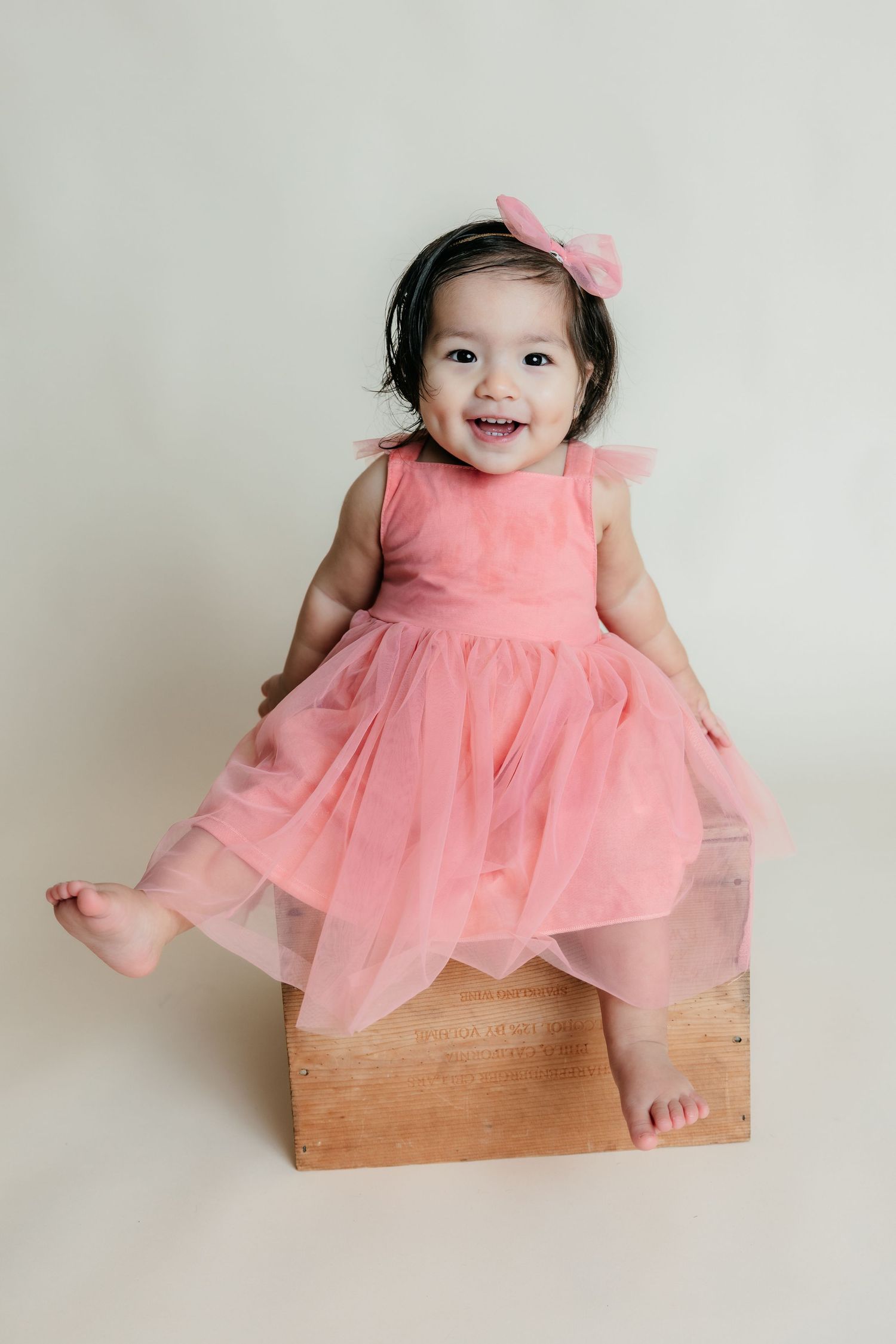 Happy baby girl in a pink tulle dress with a bow headband sitting on a wooden box during a milestone photoshoot.