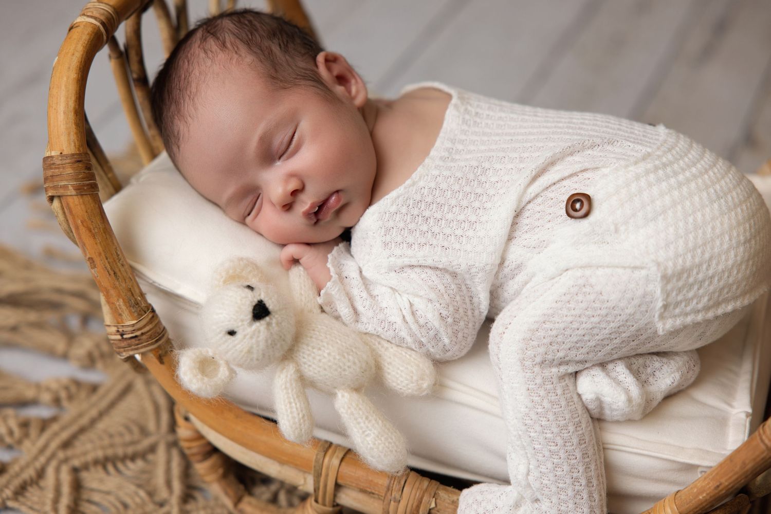 A peaceful newborn baby sleeps in a wicker basket wearing a white knit romper beside a small teddy bear.