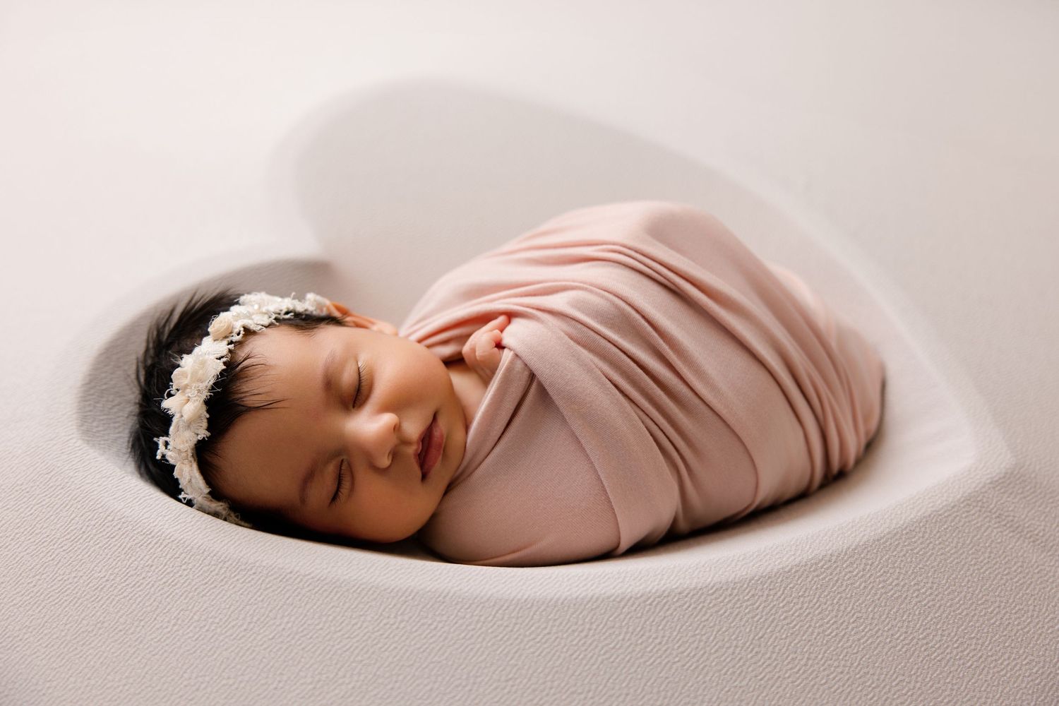 Sleeping newborn baby wrapped in pink swaddle with floral headband resting in heart-shaped white bowl.