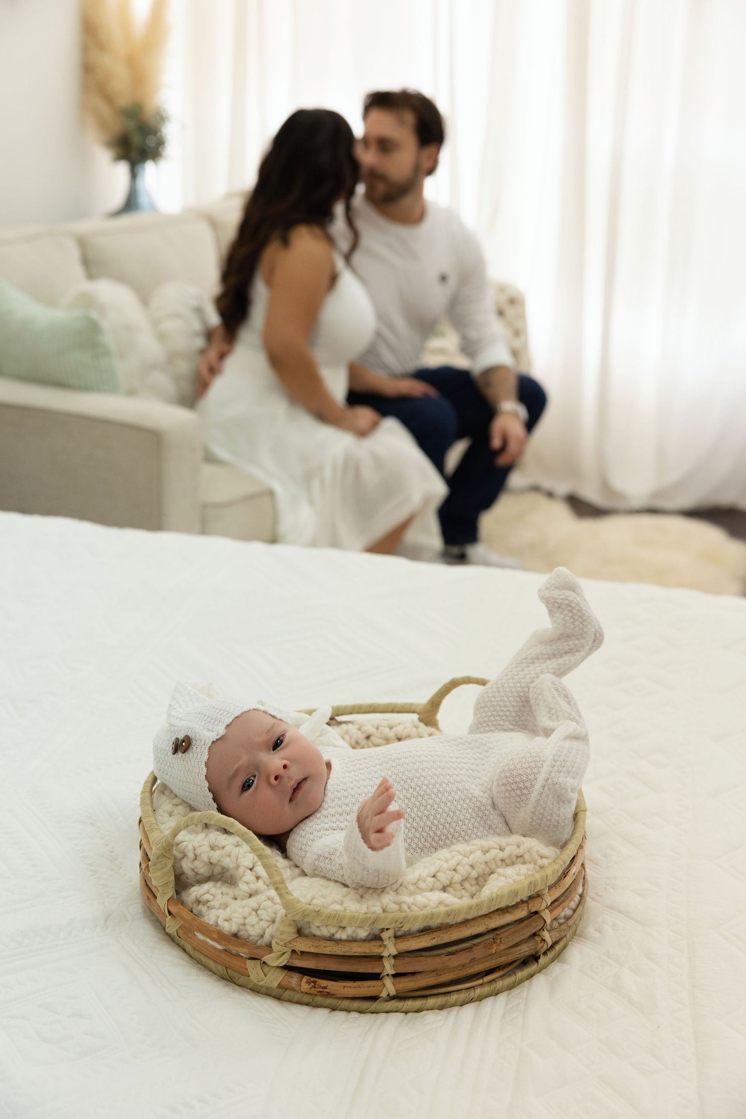 Newborn baby in white knit outfit lying in wicker basket on bed with parents sharing a tender moment in background.