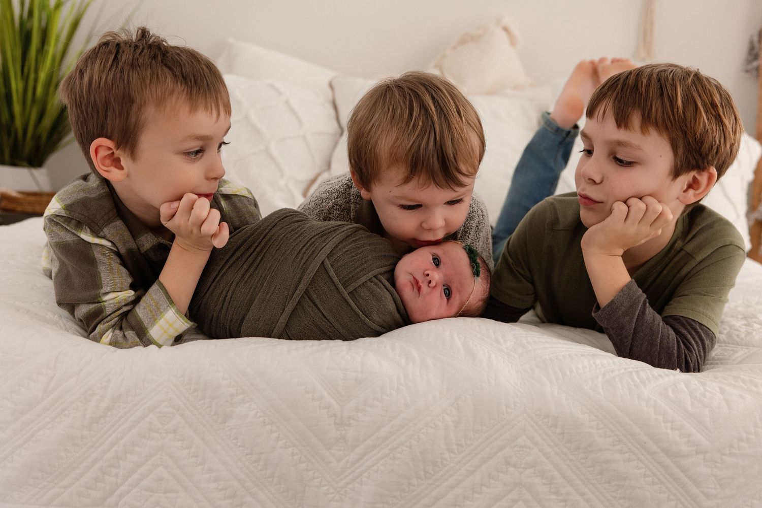 Three brothers lying on a white bed, two older boys lovingly gazing at their newborn sibling wrapped in a green swaddle.