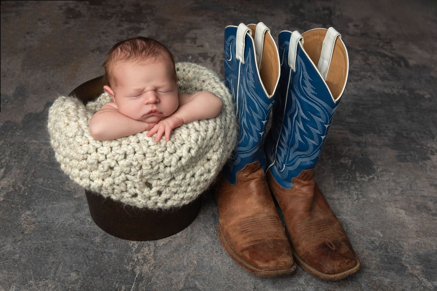 Newborn baby sleeping in a rustic bucket wrapped in cream knit blanket beside blue cowboy boots.