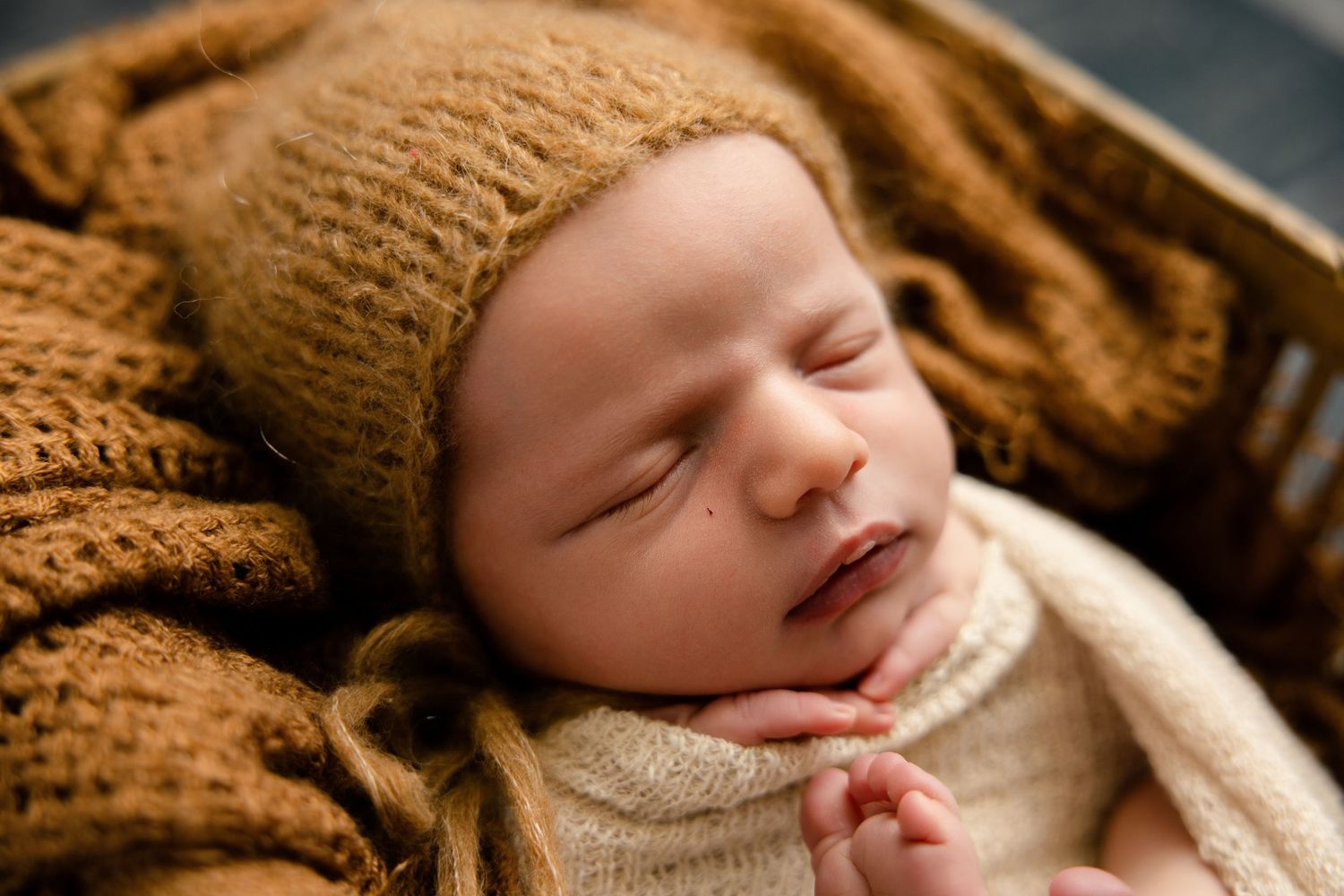 Sleeping newborn baby in a knit bonnet and cream wrap, nestled in a cozy brown basket for a studio photo.
