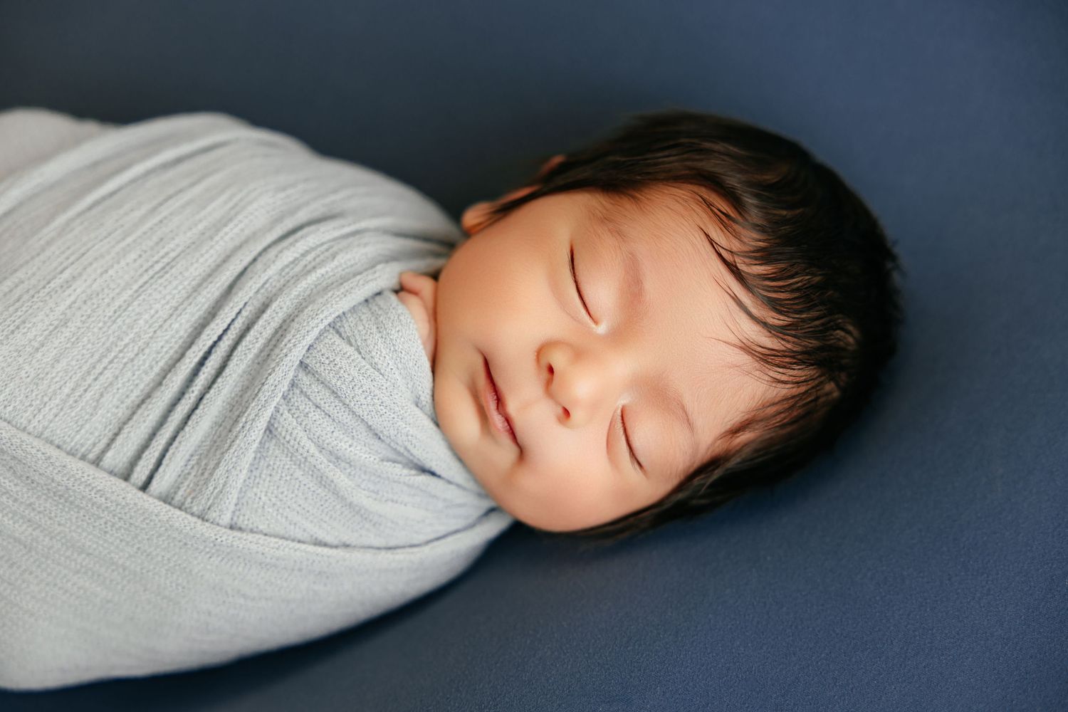 Peaceful newborn baby swaddled in a soft gray wrap, sleeping on a dark blue background.