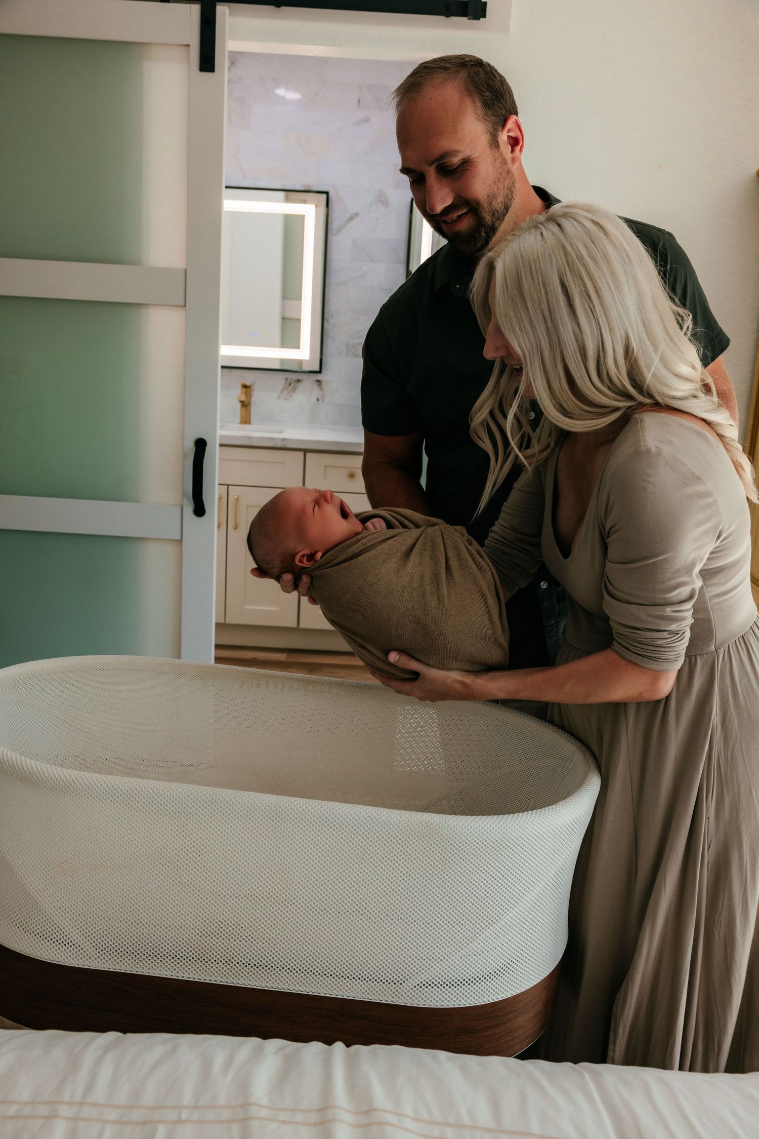Parents lovingly place their swaddled newborn into a white bassinet in a softly lit nursery.