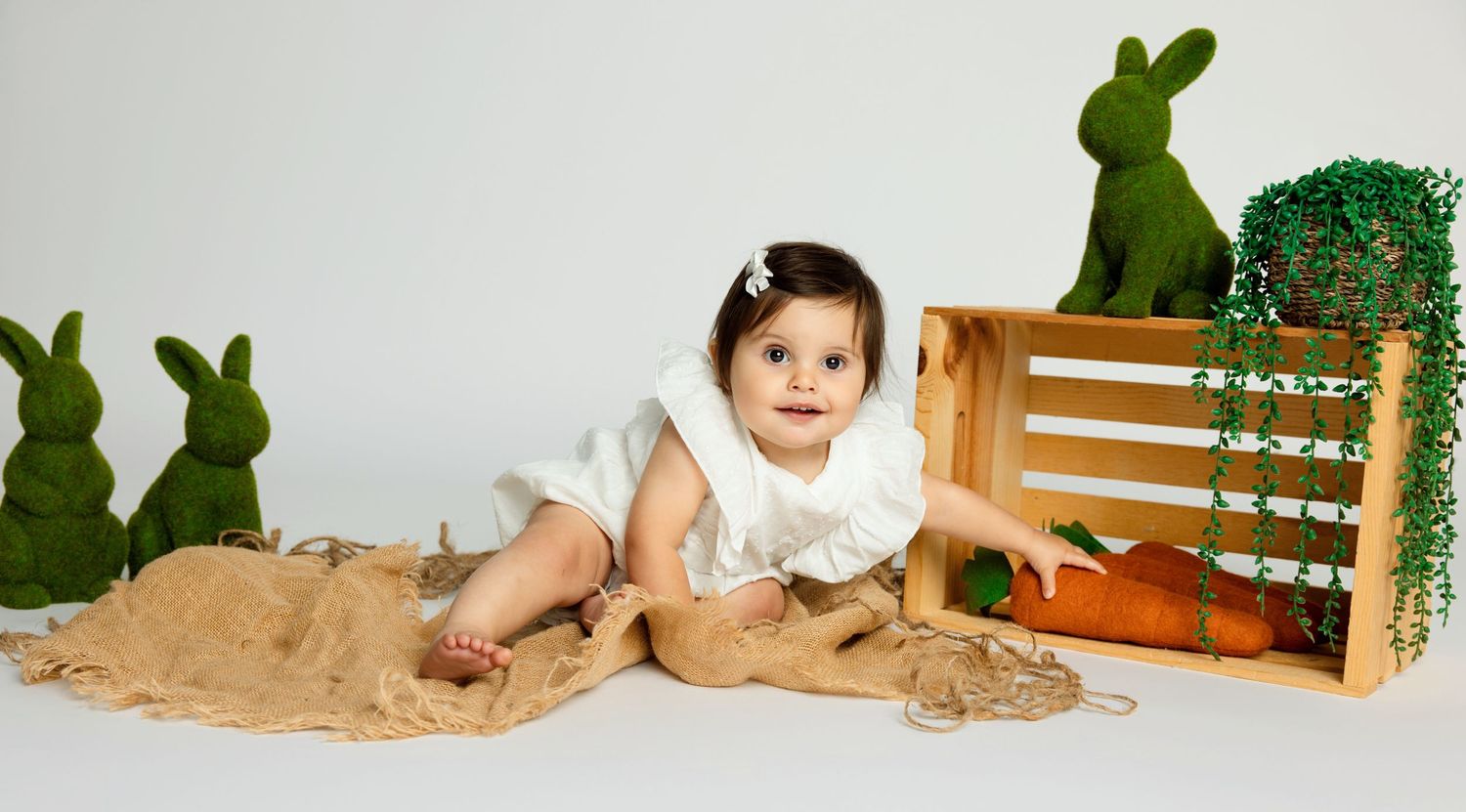 Adorable baby girl in white dress sitting with green bunny decorations and wooden crate in Easter photo session. Fallbrook Newborn Photograp