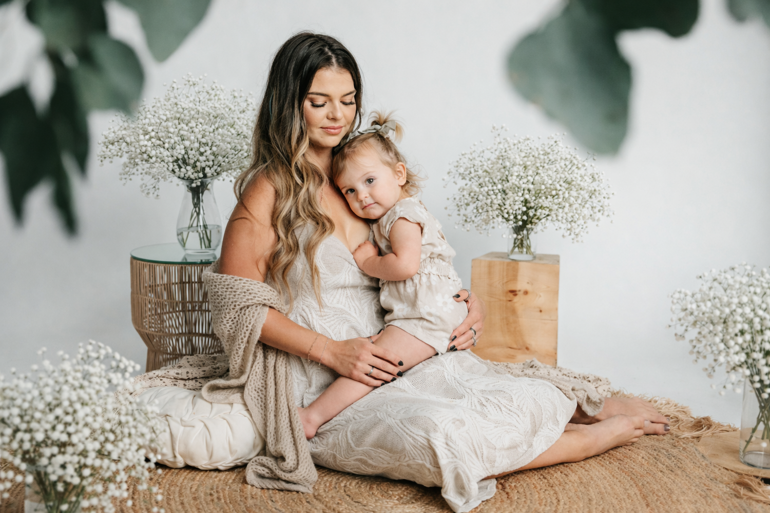 A mother in a white dress lovingly embraces her toddler daughter surrounded by white flowers in a studio.