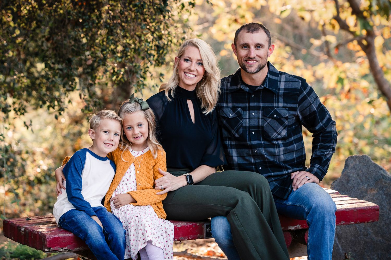 Family of four smiling together on a red bench with colorful autumn foliage in the background.
