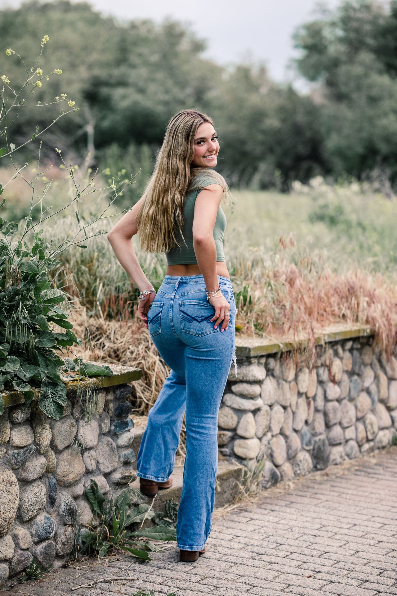 Smiling blonde woman in green crop top and flare jeans posing along stone wall in outdoor nature setting.