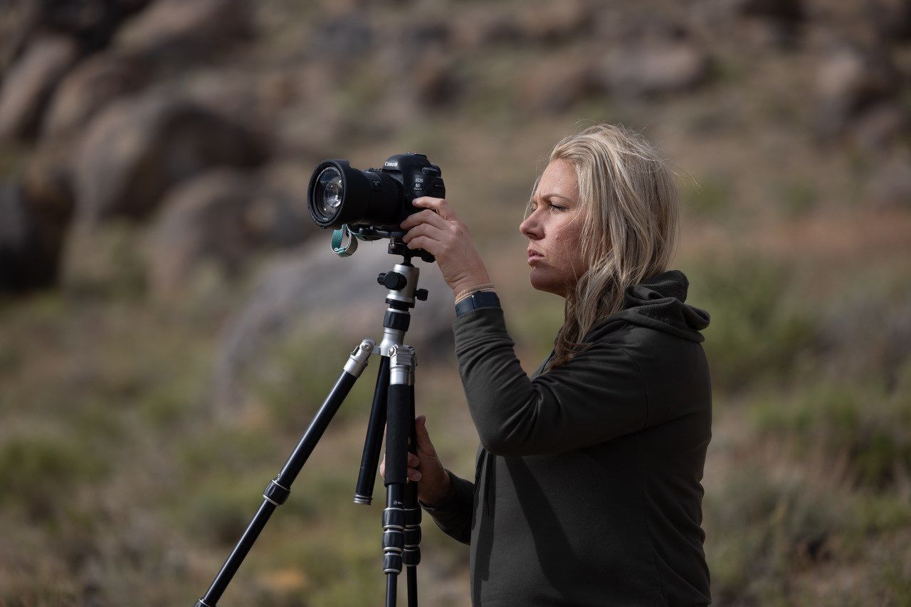 A travel teaching photographer with a camera and tripod captures scenic landscape views in a mountainous desert terrain.