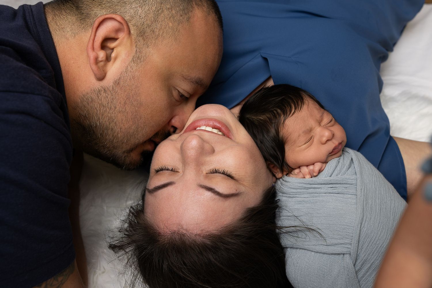 A couple lies in bed smiling, the man kissing the woman's cheek as they cuddle their sleeping newborn baby.