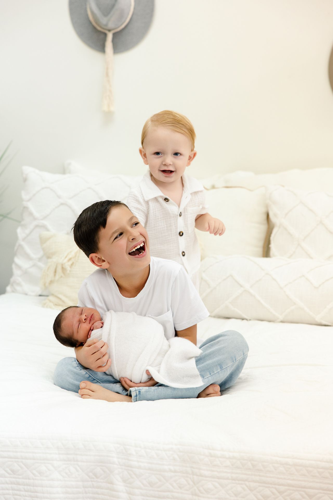 Three brothers pose on a white bed, older boy holds swaddled newborn while toddler stands smiling behind them.