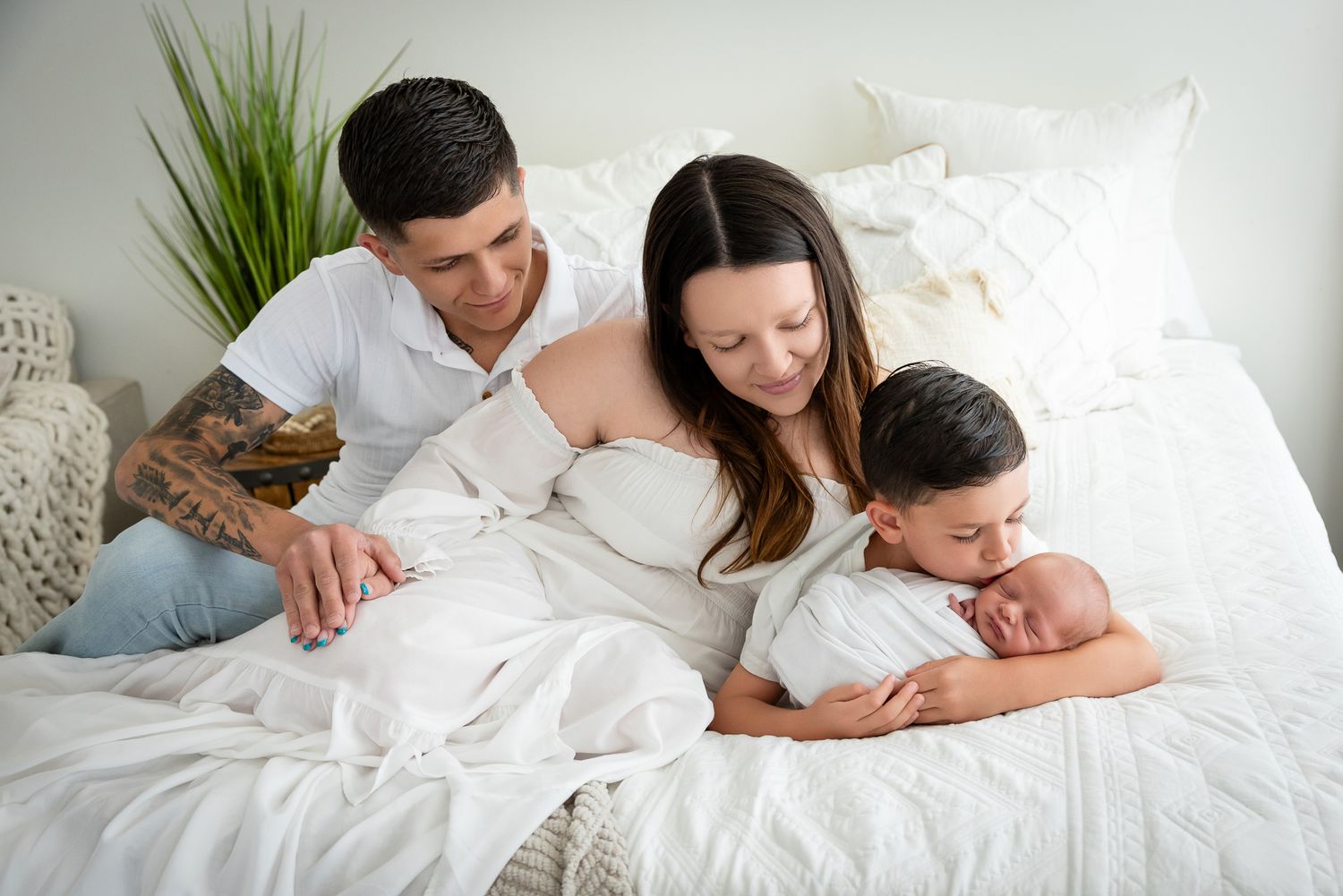 Family of four on white bed, parents with tattooed dad, older son, and newborn baby in cozy bedroom setting.