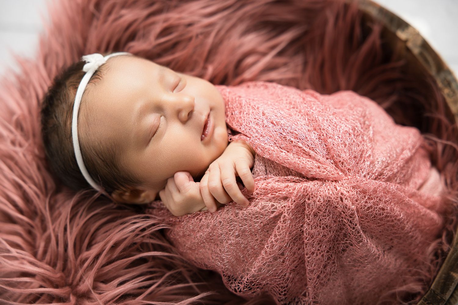 Sleeping newborn baby girl wrapped in pink knit swaddle with white bow headband in a cozy fur-lined basket.