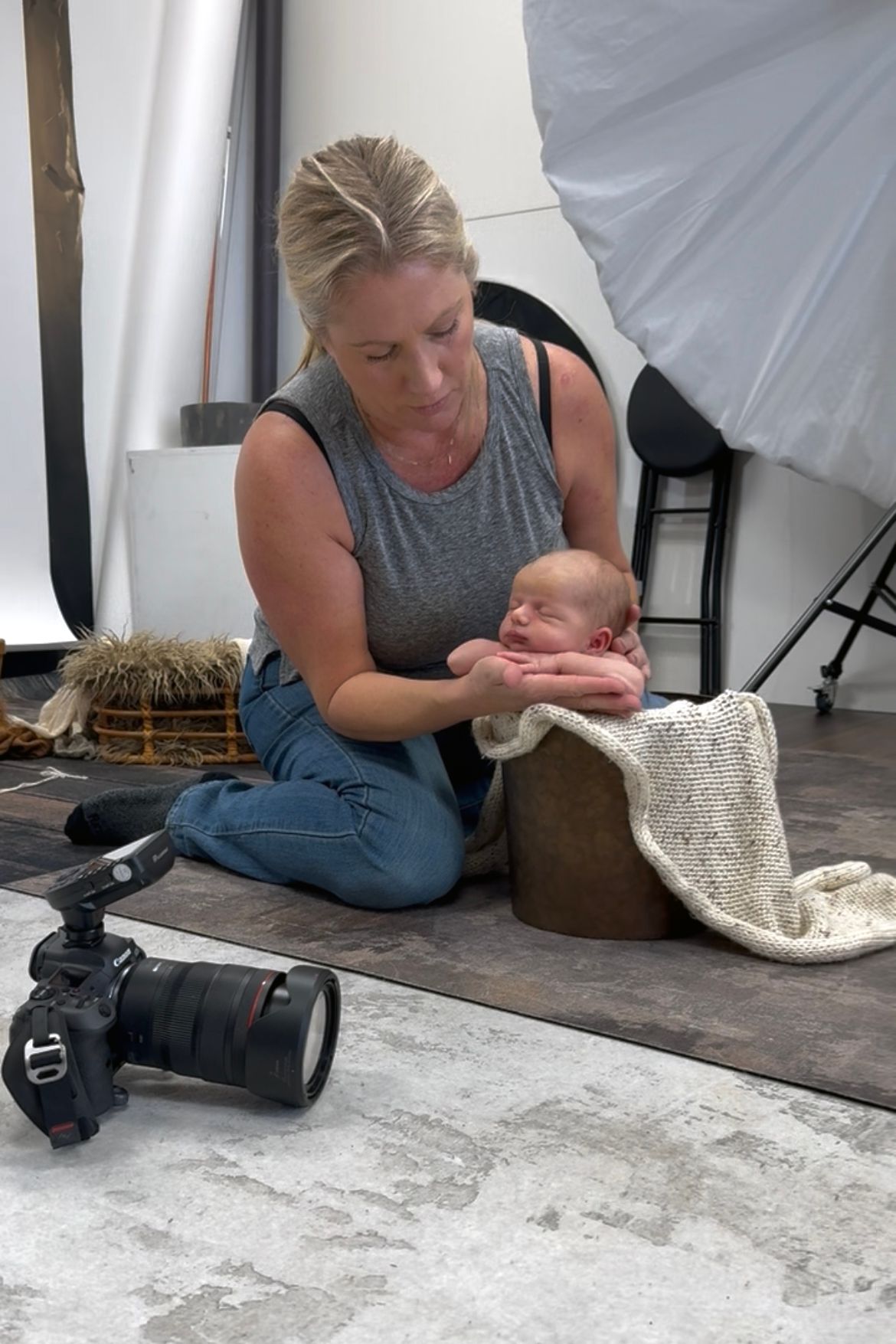 Photographer poses newborn baby in wooden bucket with knit blanket during studio photo session.