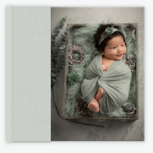 White leather photo album featuring a smiling newborn baby girl swaddled in green wrap surrounded by flowers. Vista Newborn Photographer