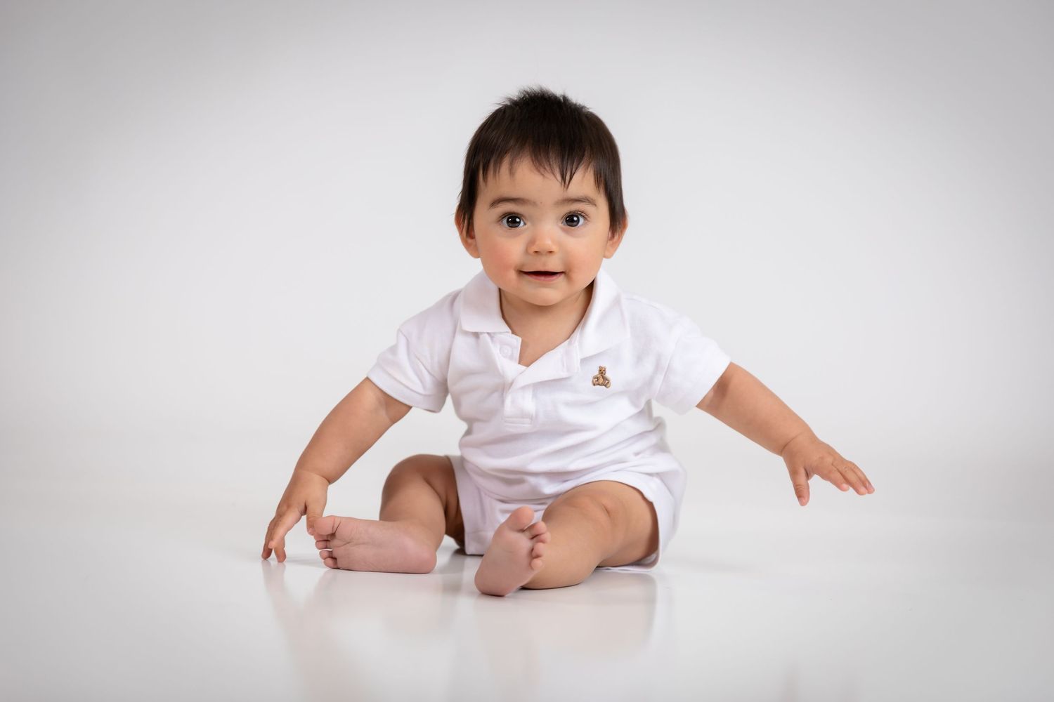 Adorable baby in white polo onesie sitting on white floor, smiling at camera during professional photo shoot.
