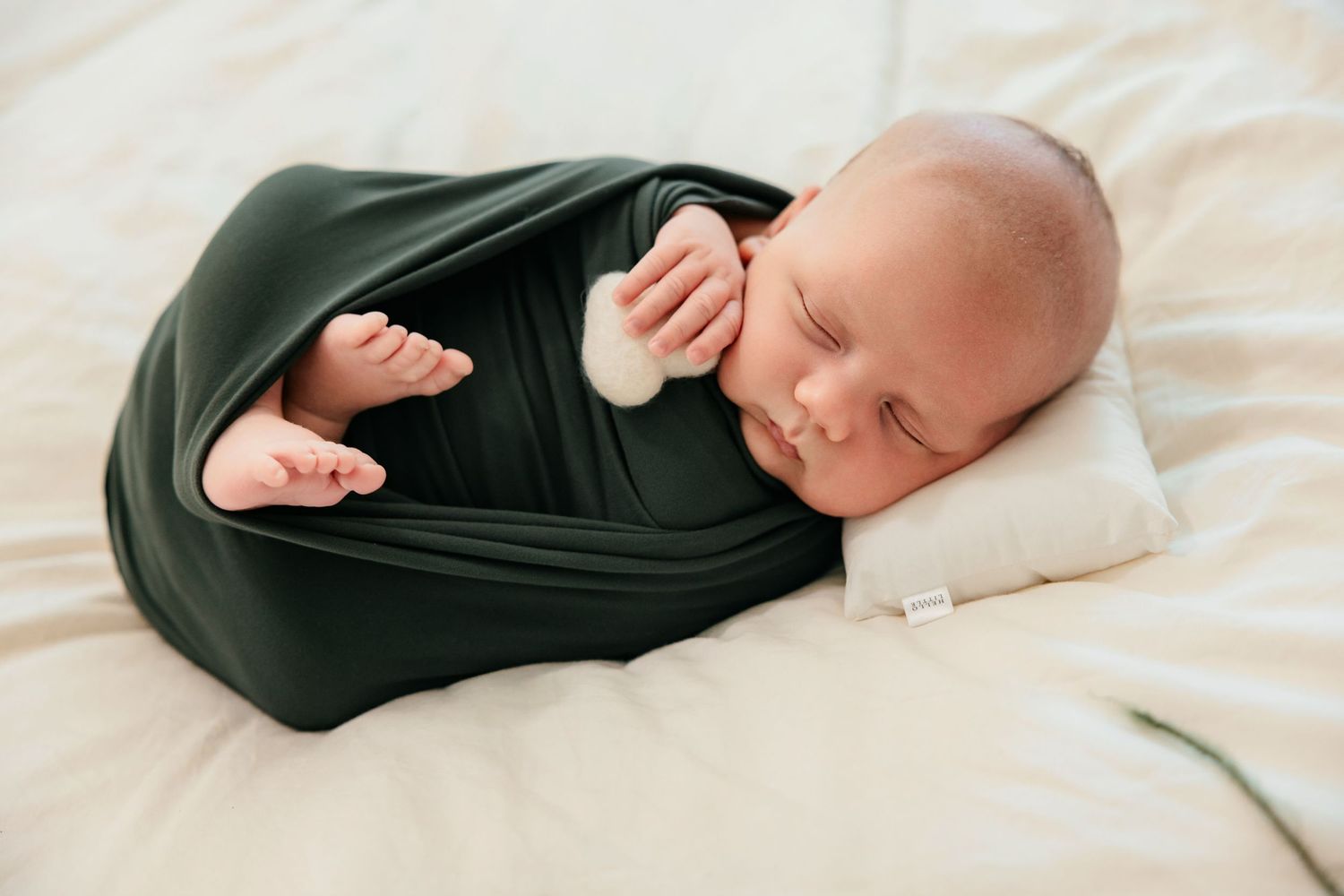 Newborn baby swaddled in dark green wrap sleeping peacefully on white bedding with tiny feet peeking out.