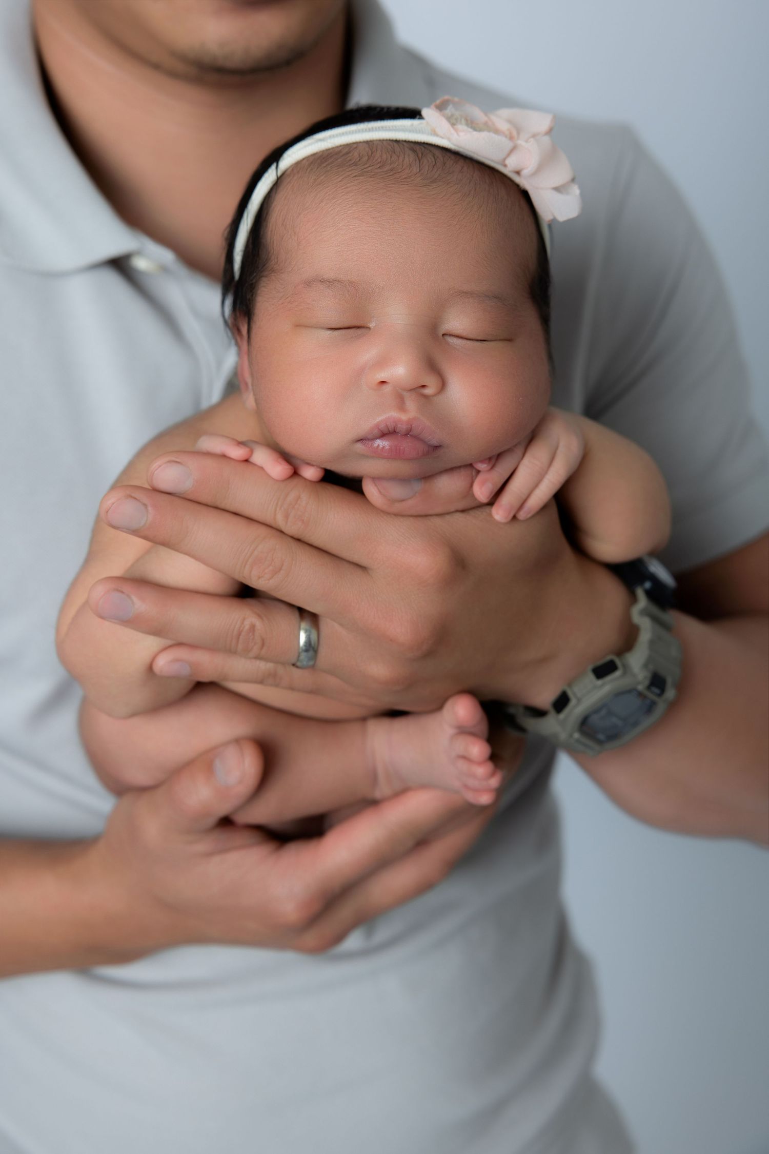 Father gently cradling sleeping newborn baby girl wearing pink floral headband in his hands.