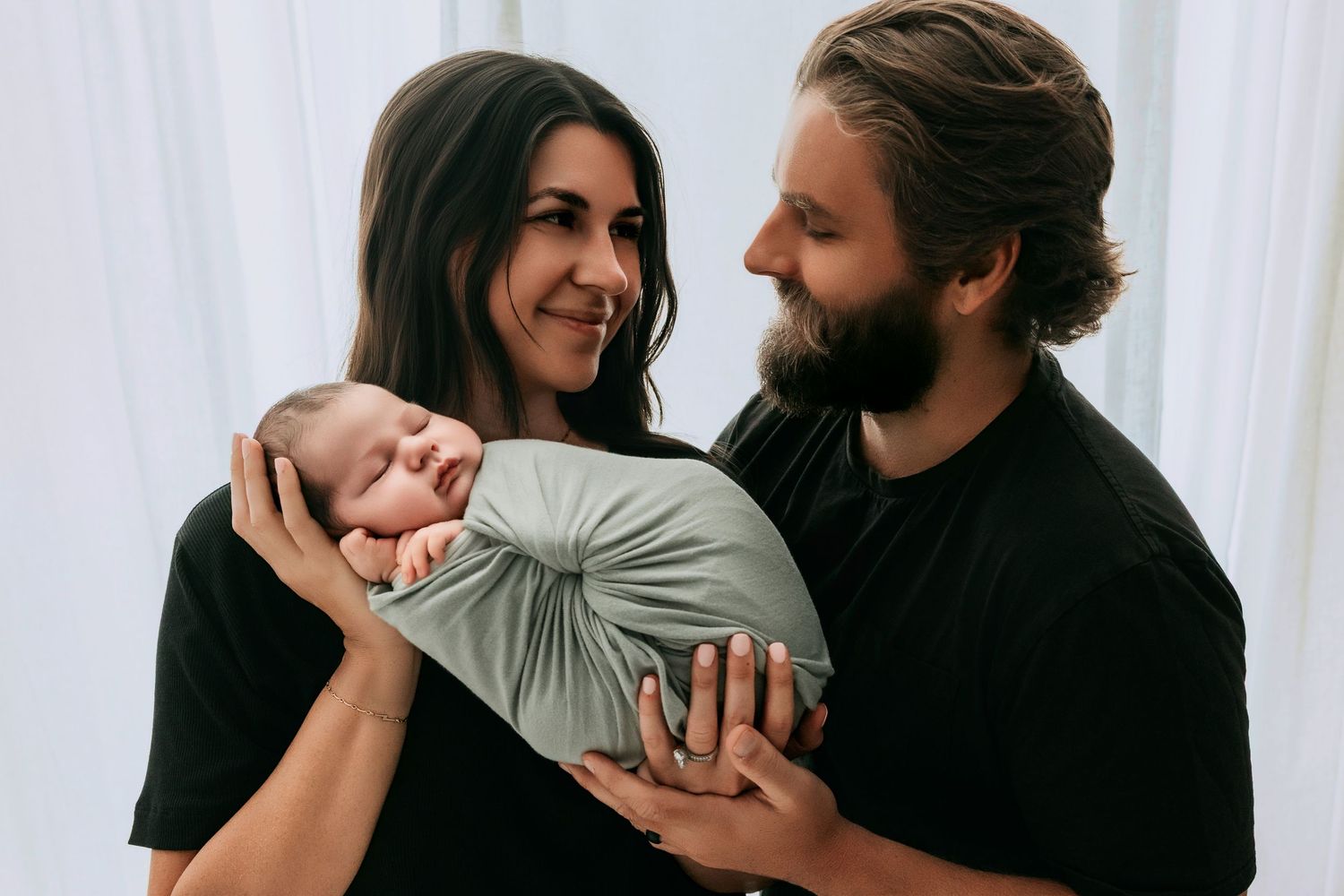 Happy parents in black shirts holding their swaddled newborn baby in a soft gray wrap.