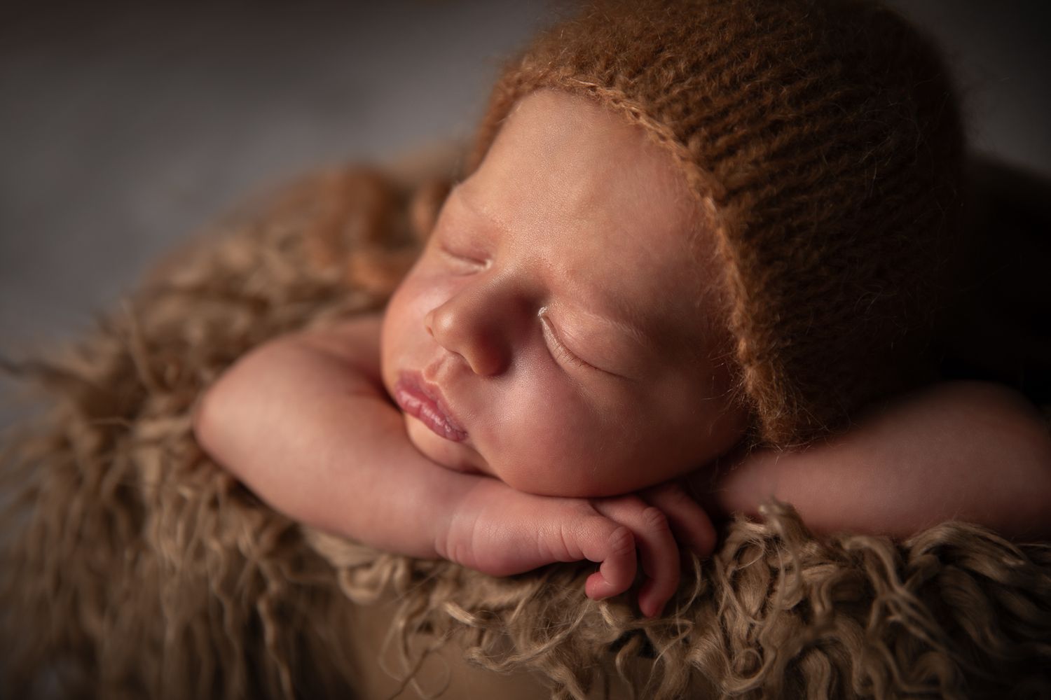 Sleeping newborn baby wearing a brown knit bonnet resting on a fluffy textured fur surface.
