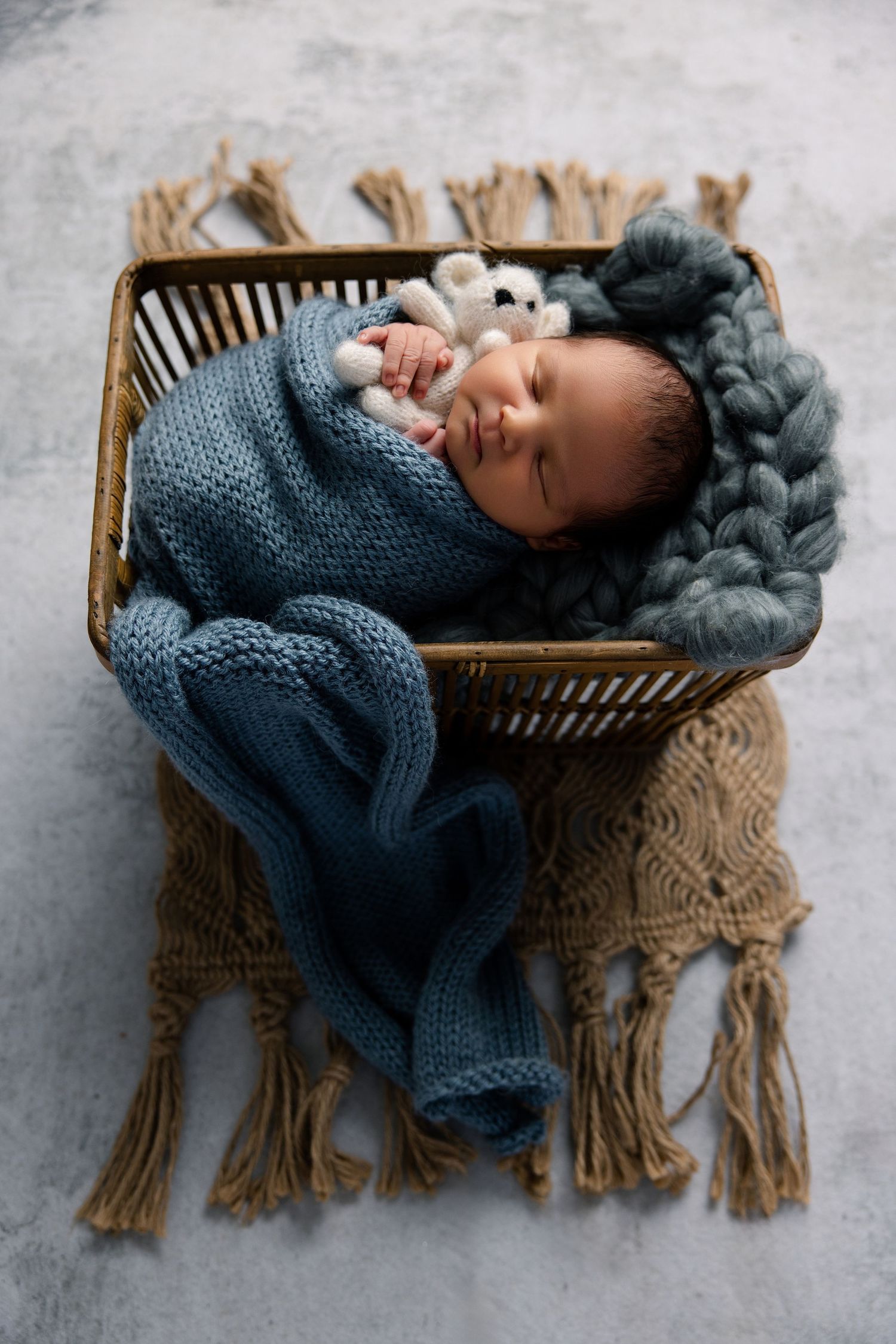 Peaceful newborn baby swaddled in blue knit wrap, sleeping in rustic wicker basket with chunky yarn blanket and tiny teddy bear.
