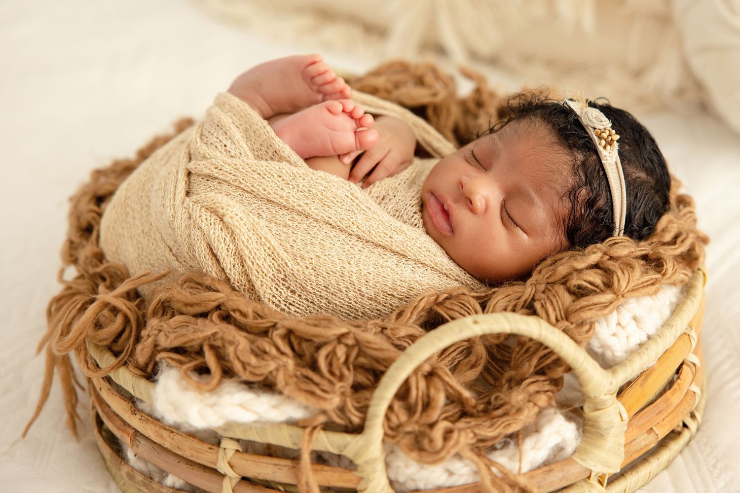 Peaceful newborn wrapped in tan swaddle with gold headband, resting in a wicker basket with brown yarn.
